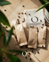Coffee bean sample packs with Coffee Stage logo on wooden surface, scattered coffee beans, and green plant leaves in foreground, representing a premium coffee tasting experience