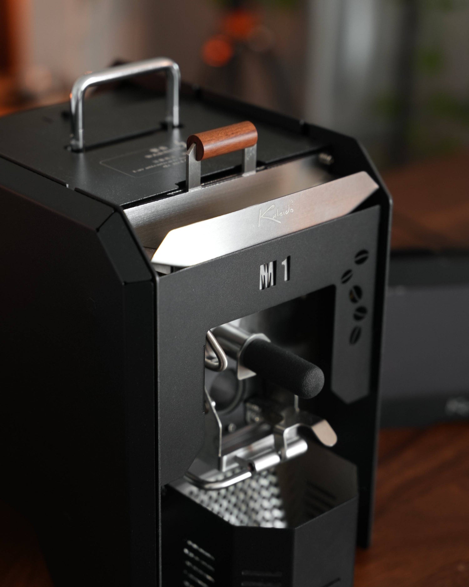 KALEIDO M1 coffee roaster machine, black compact design with metal accents, featuring a front viewing window and side handle, showcasing professional-grade home roasting capabilities