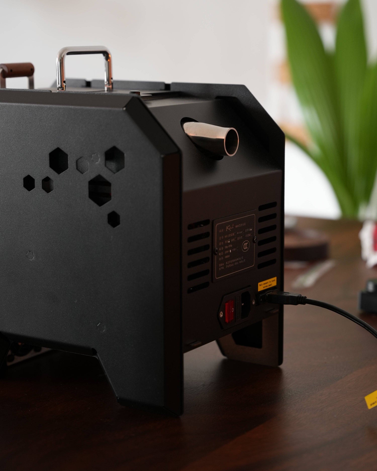 KALEIDO M1 coffee roaster machine with sleek black design, hexagonal vents, and copper handle, displayed on wooden surface with plant in background