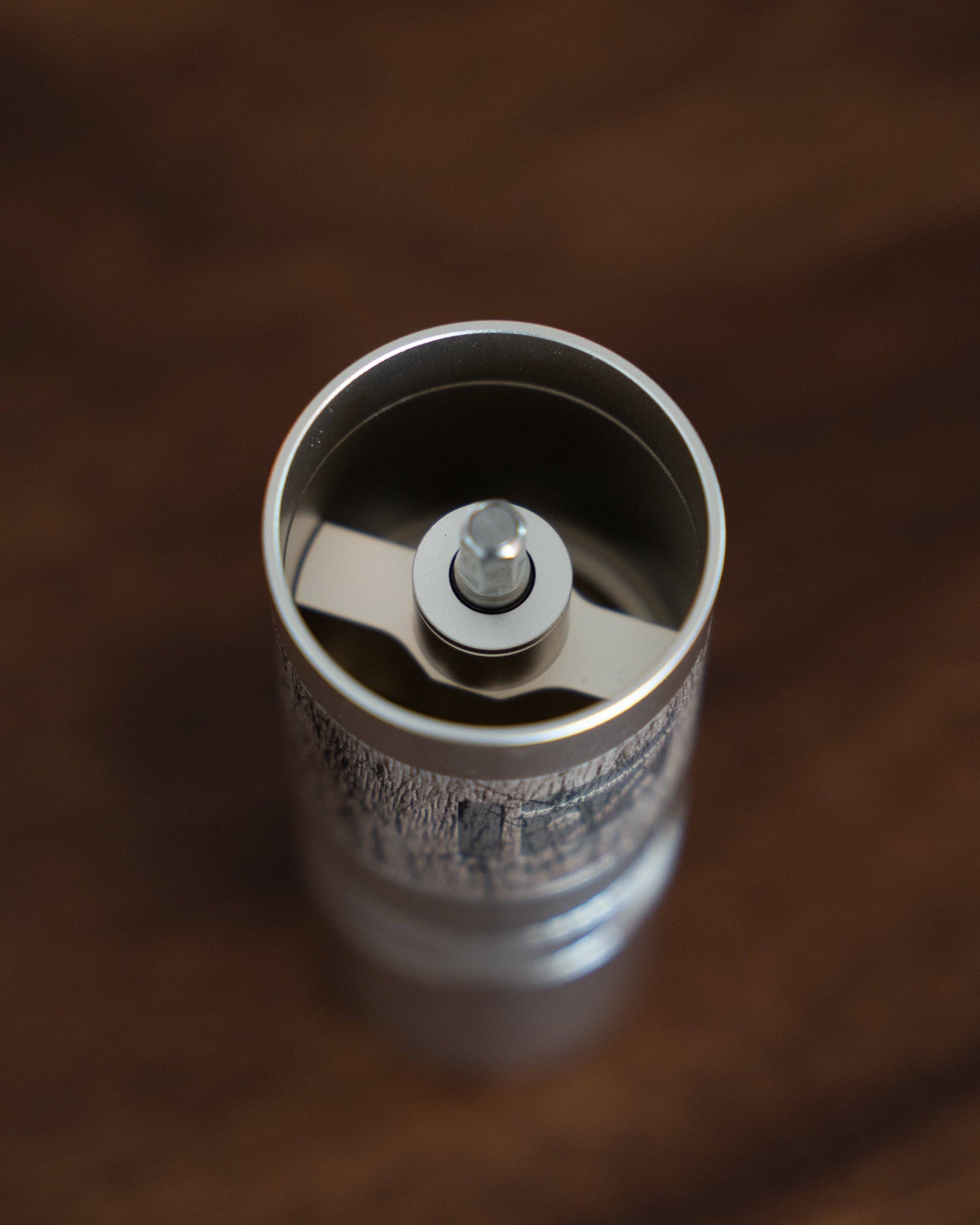 Top view of 1Zpresso QS2 manual coffee grinder, showing stainless steel burr set inside compact cylindrical body on dark wooden surface, highlighting precision engineering and portable design for coffee enthusiasts