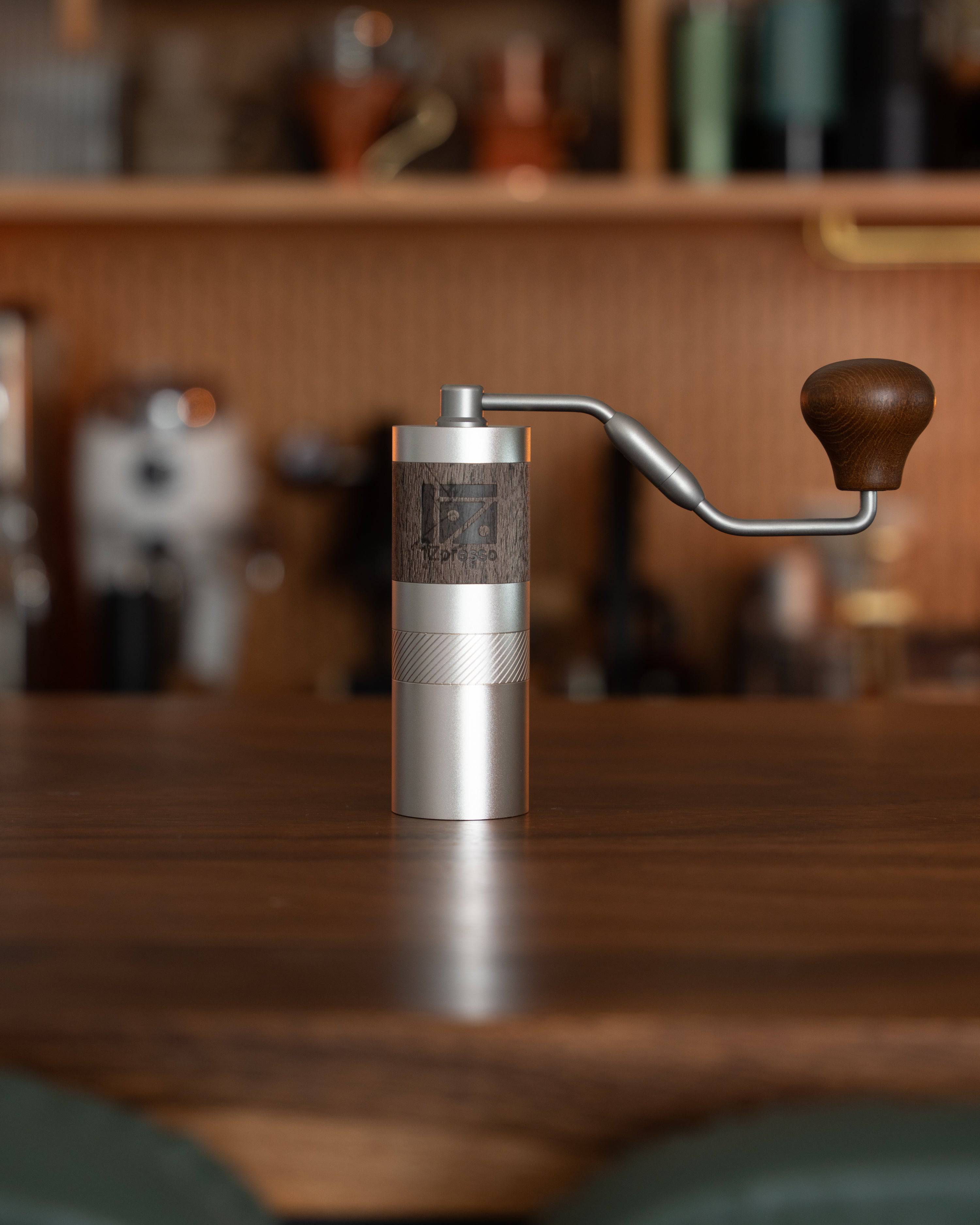 Compact 1Zpresso QS2 manual coffee grinder with wooden handle on wooden table, surrounded by blurred coffee equipment in background