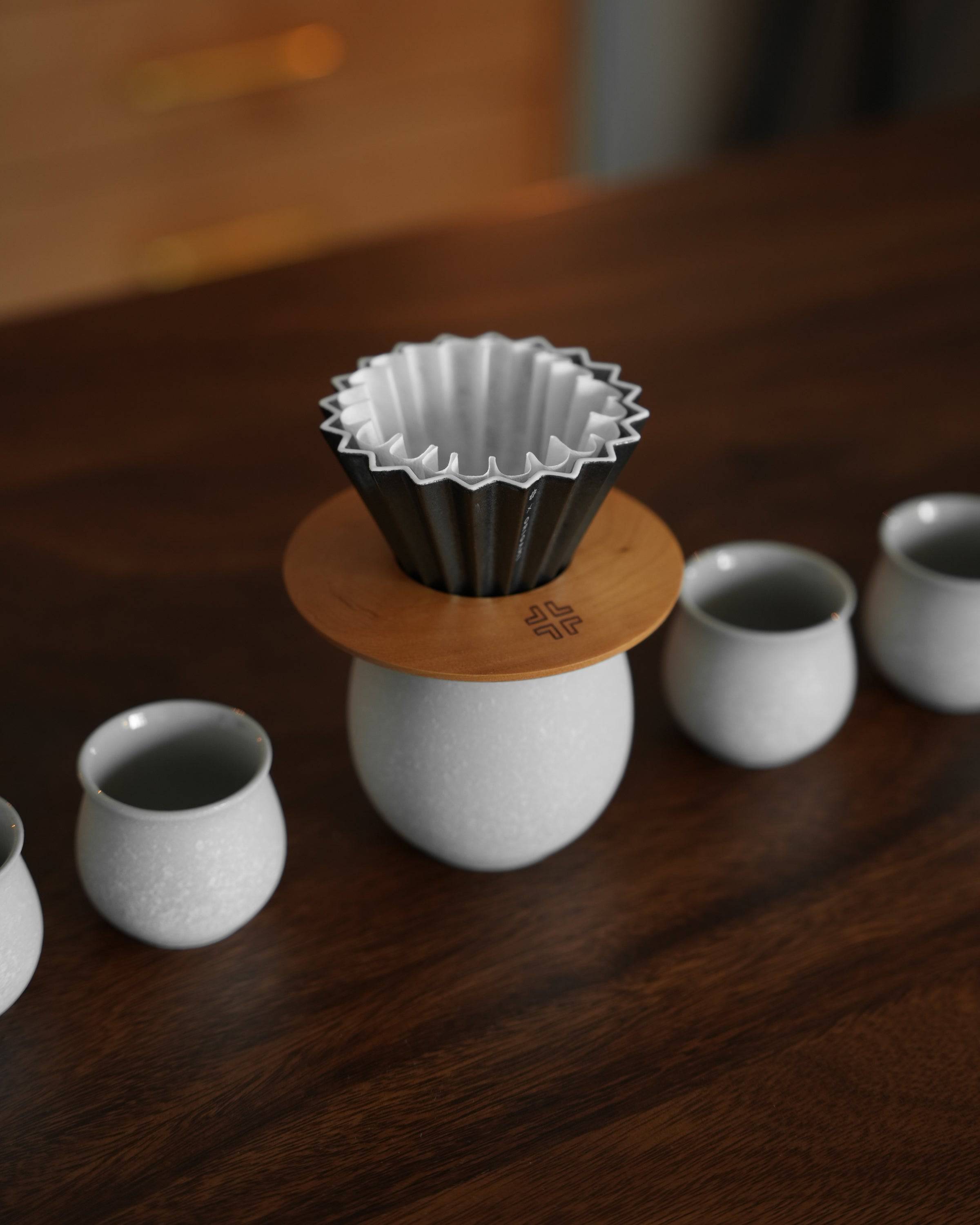 Ninety Plus Coffee Kone Ring 10.1 walnut wood stand with metal filter cone on white ceramic cup, surrounded by smaller cups on dark wooden surface, showcasing elegant coffee brewing setup