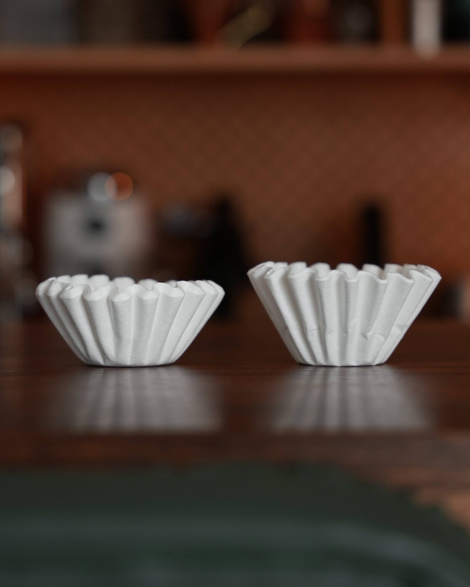 Two white, fluted coffee filter papers side by side on a dark wooden surface, showcasing the unique shape and design of Ninety Plus Coffee Kone Filter 10.1 for premium coffee brewing.