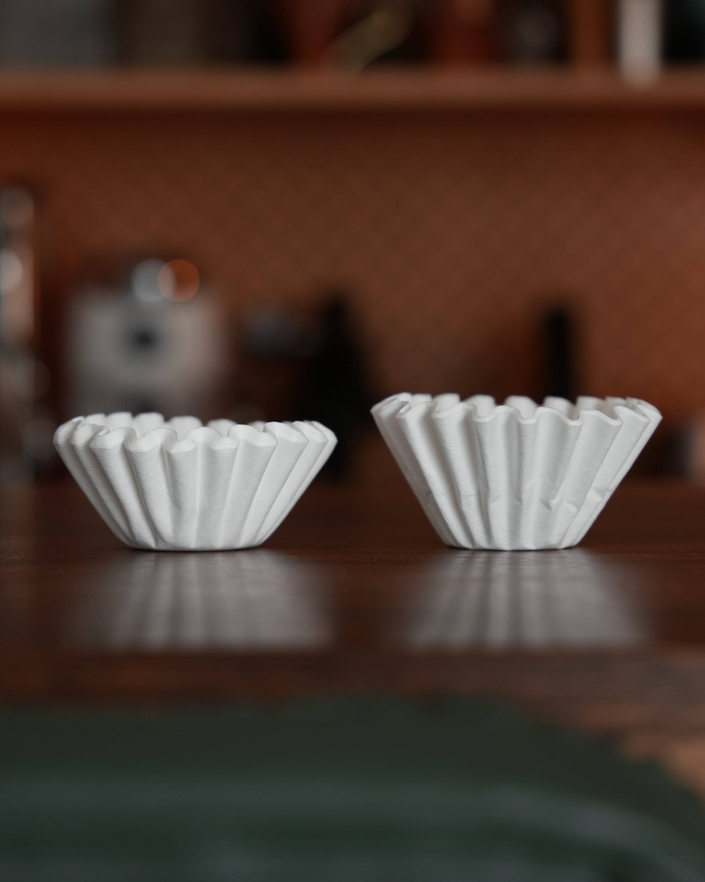 Two white, fluted coffee filter papers side by side on a dark wooden surface, showcasing the unique shape and design of Ninety Plus Coffee Kone Filter 10.1 for premium coffee brewing.
