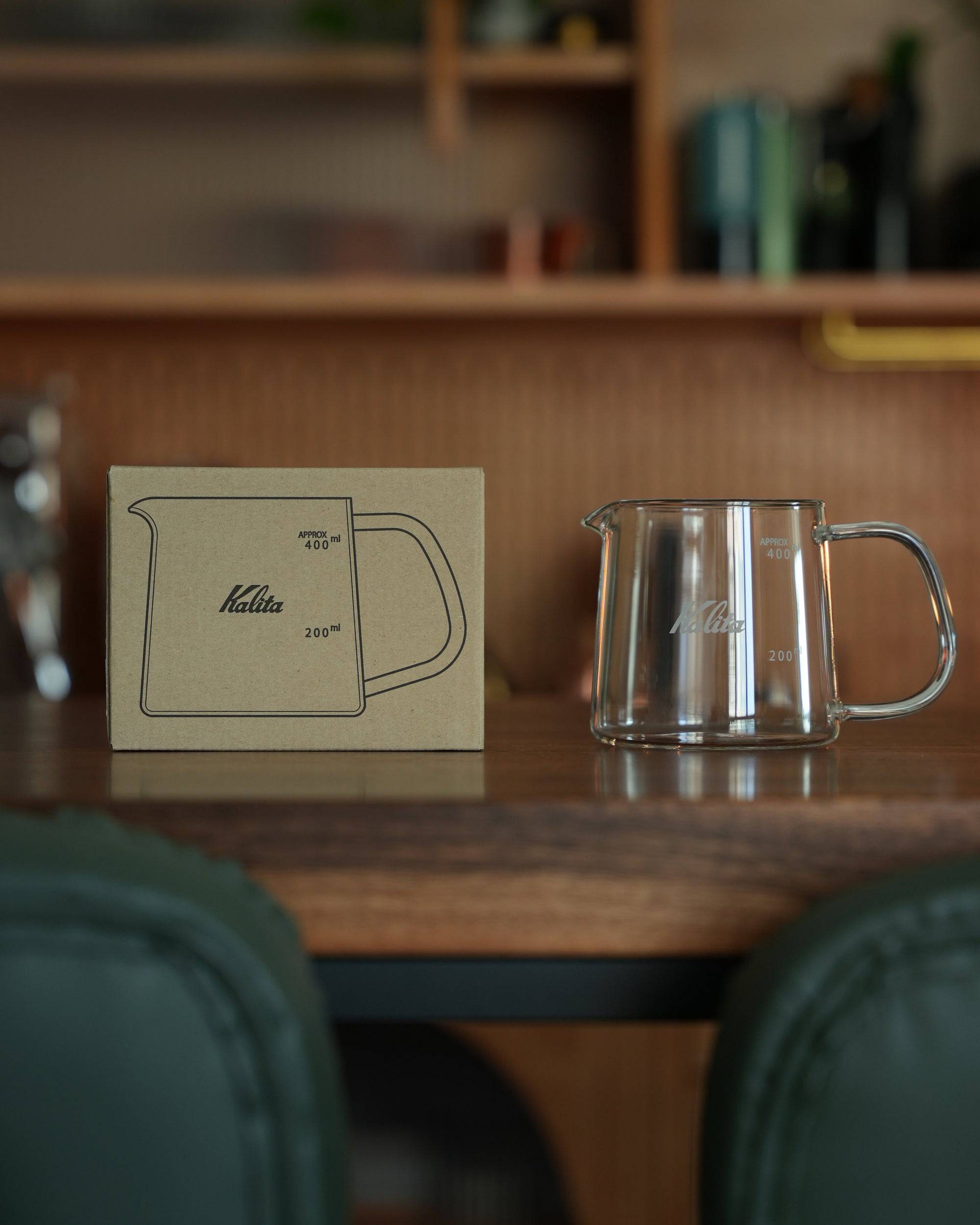 Kalita JUG400 heat-resistant glass coffee jug displayed on wooden table with product box, showcasing its elegant design, clear measurement markings, and comfortable handle for perfect pour-over coffee brewing