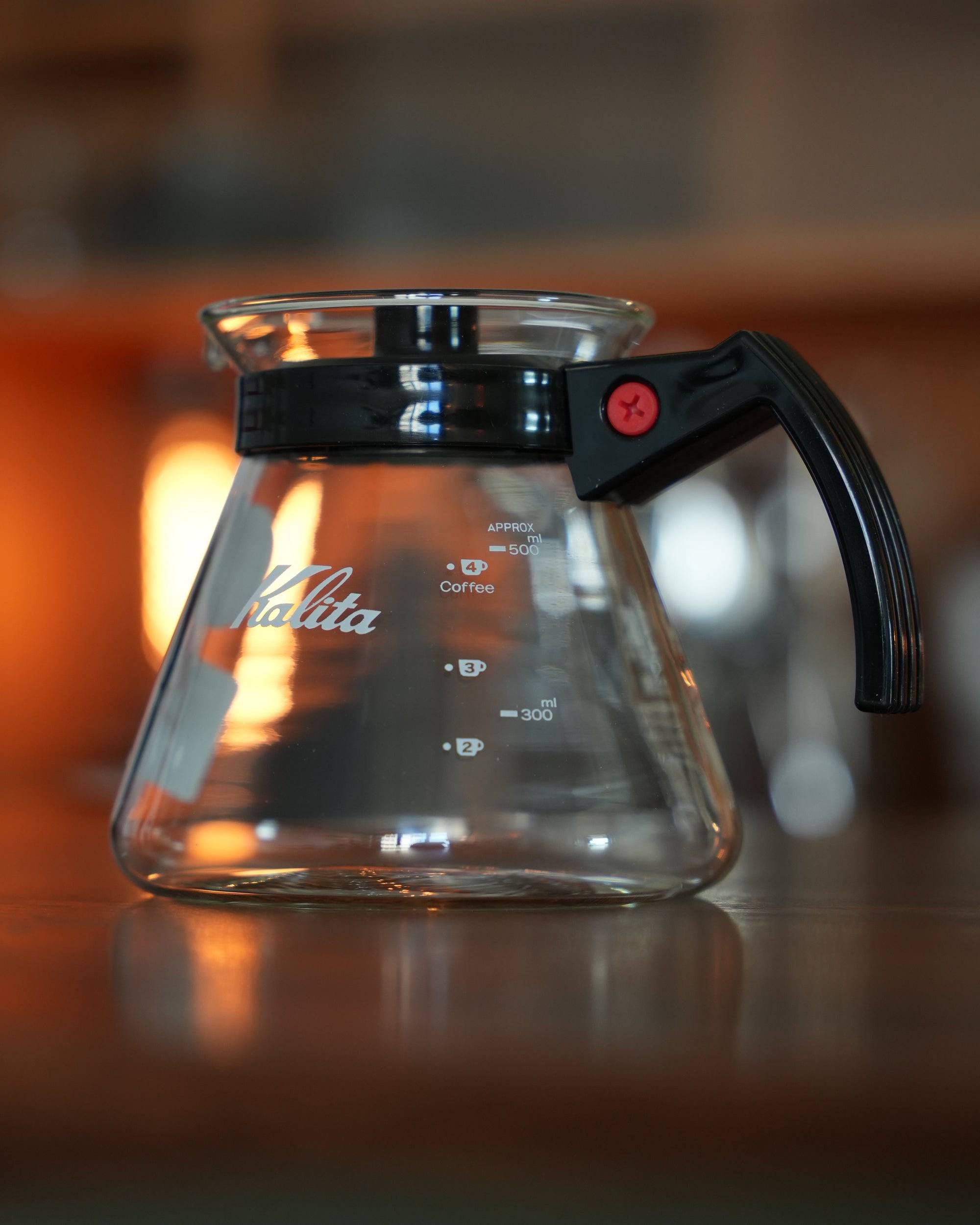 Kalita 500 Server N heat-resistant glass coffee pot in warm lighting, showing transparent body with measurement markings, Kalita logo, black handle and lid, perfect for brewing and serving coffee or tea