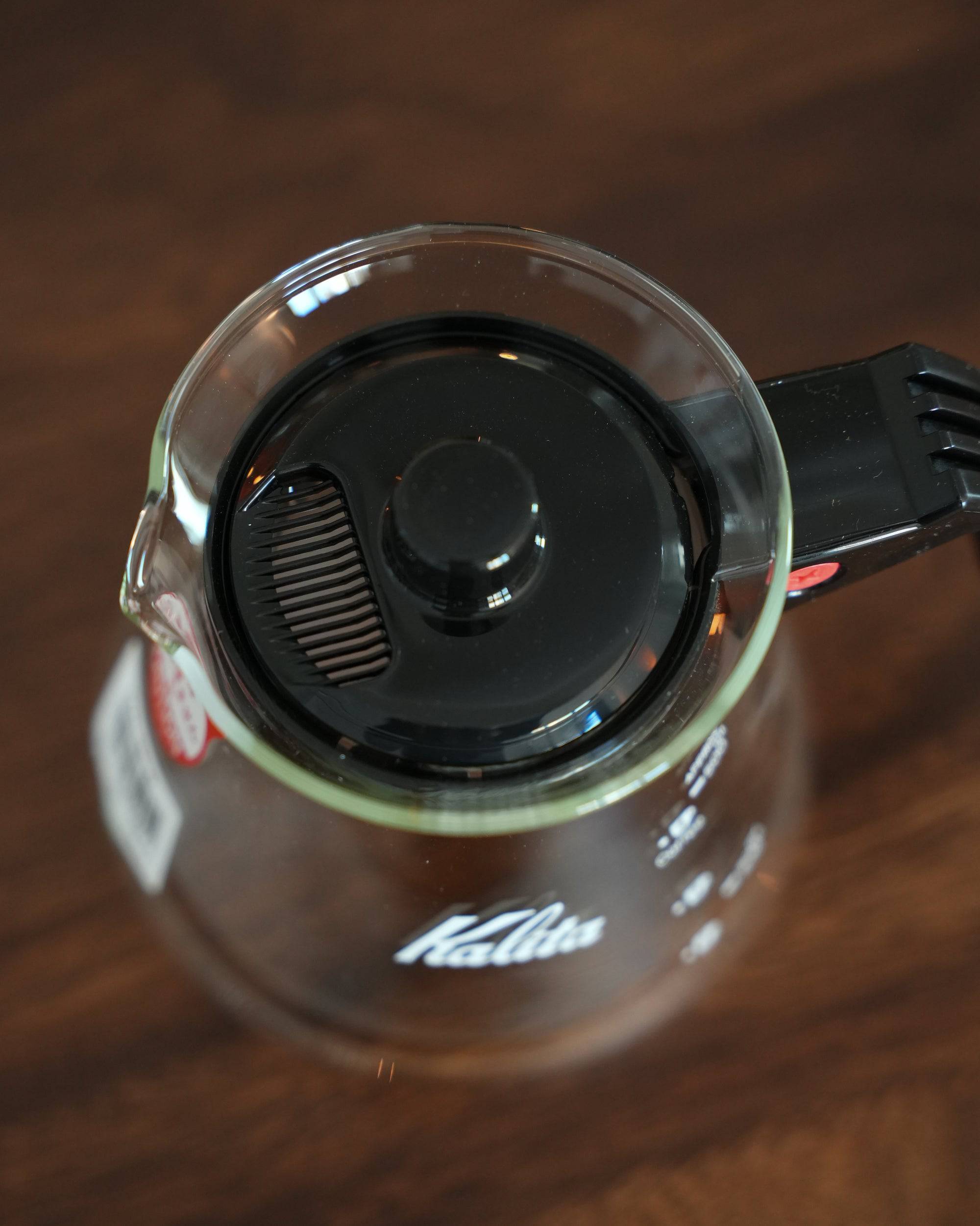 Kalita 500 Server N heat-resistant glass coffee pot with black lid and handle, viewed from above on wooden surface. Clear glass shows Kalita logo. 500ml capacity for brewing coffee or tea.