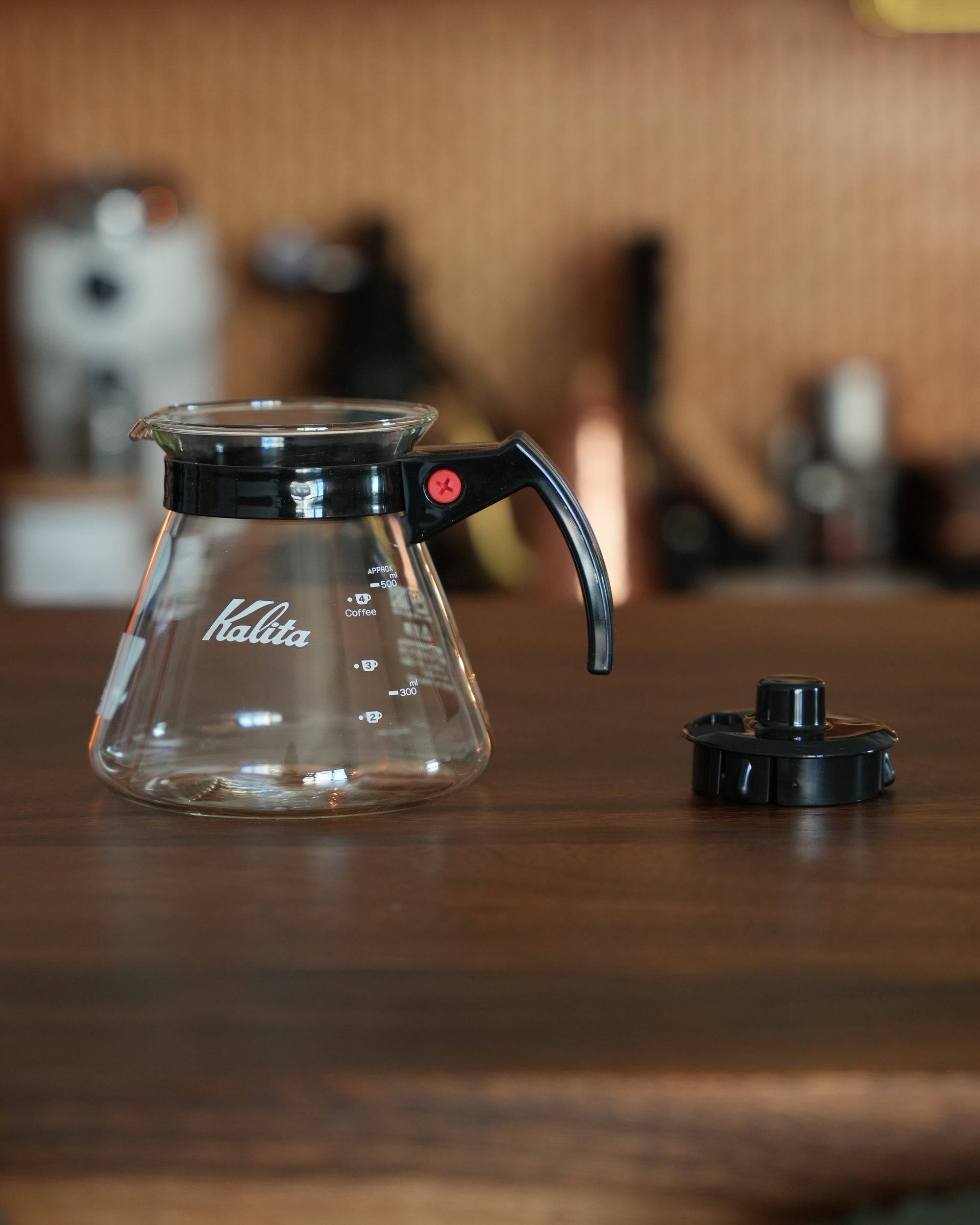 Kalita 500 Server N heat-resistant glass coffee pot with black handle and lid, 500ml capacity, displayed on wooden surface with coffee equipment in background