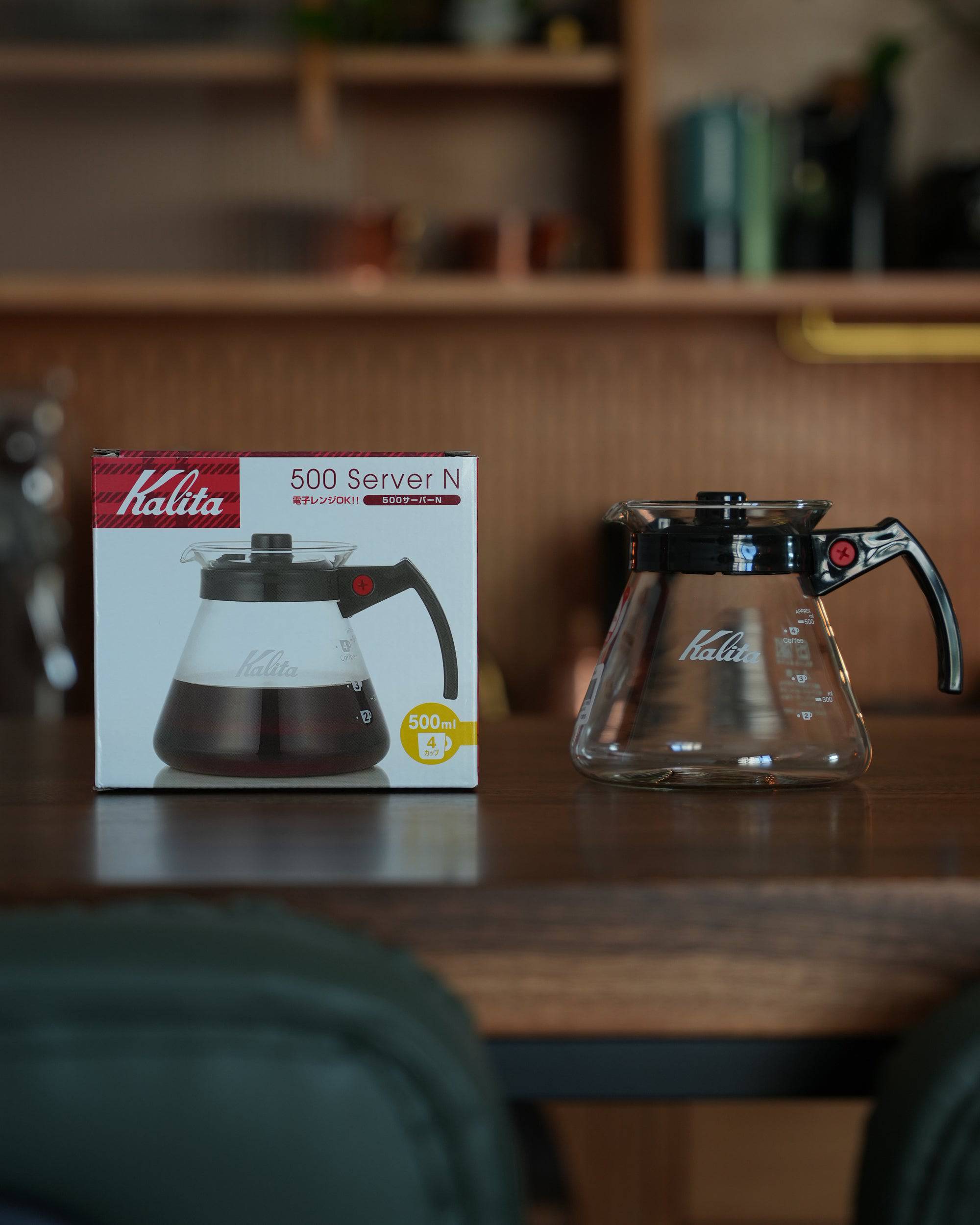 Kalita 500 Server N glass coffee pot displayed next to its product box on wooden table, showcasing transparent carafe with black handle and lid, demonstrating size and design of 500ml heat-resistant coffee server