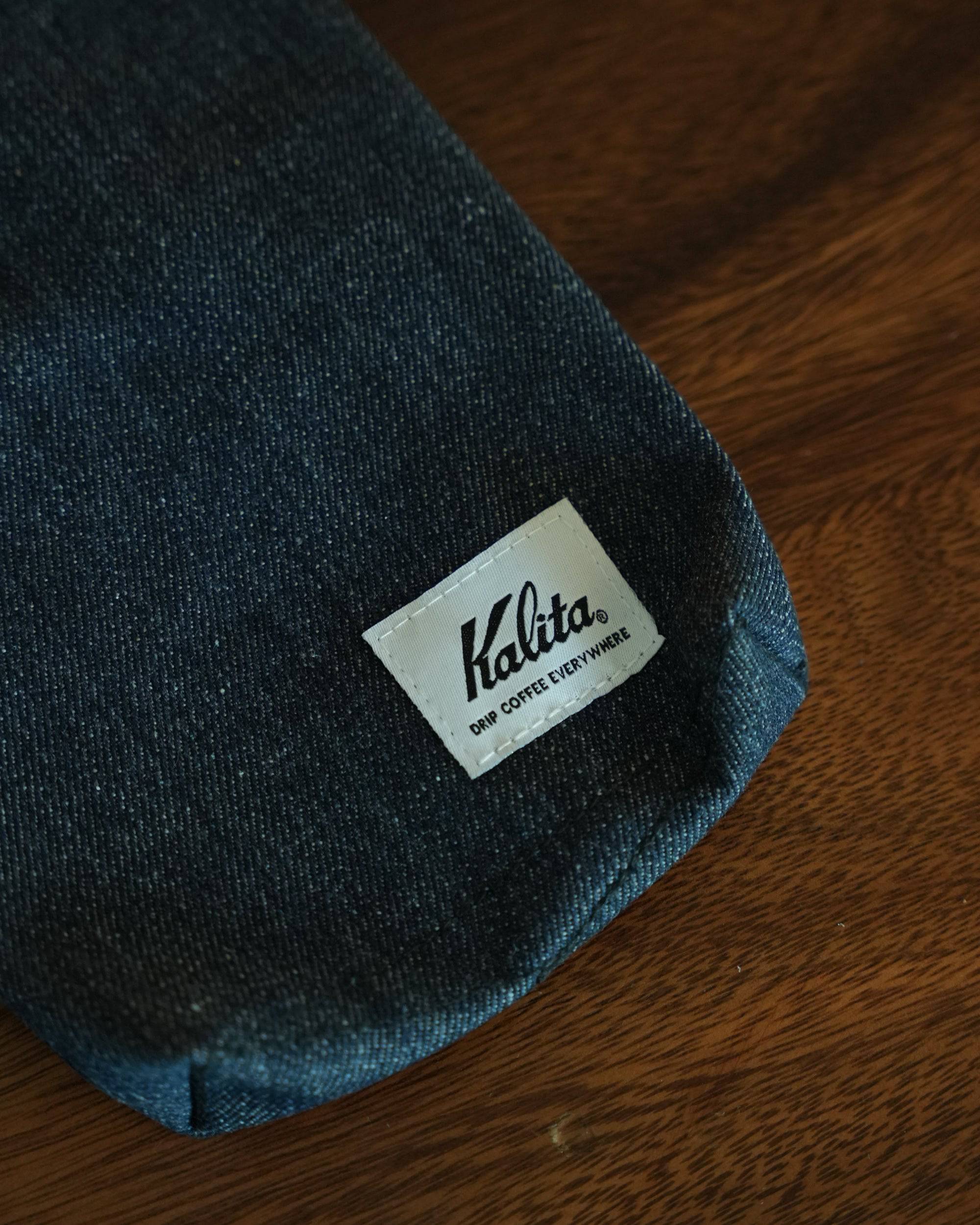 Close-up of Kalita coffee filter storage bag in dark denim fabric with white branded label, displayed on wooden surface, showcasing stylish design and quality materials for coffee enthusiasts