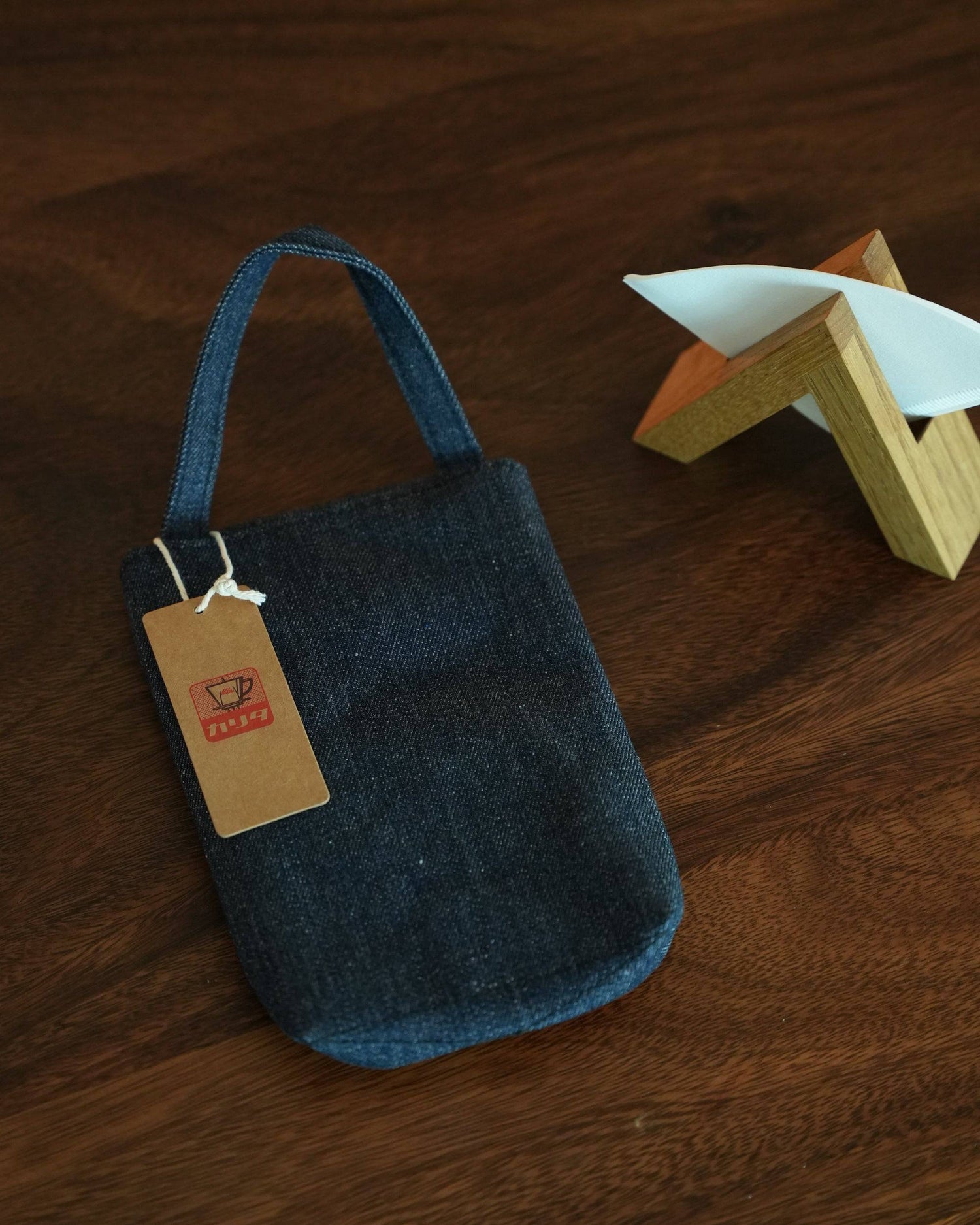 Denim Kalita coffee filter storage bag with handle and tag, next to wooden filter holder on dark wooden surface