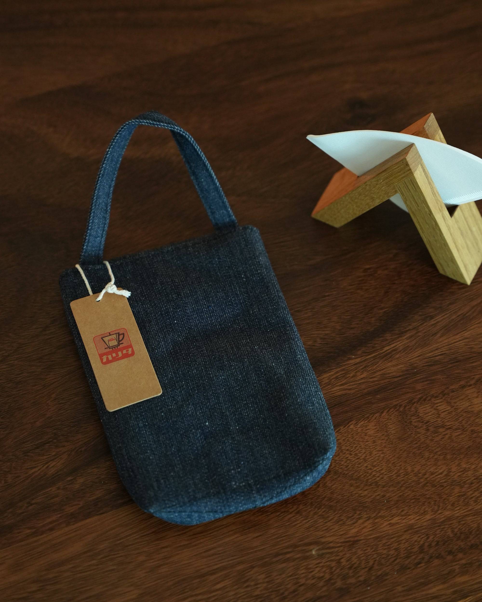 Denim Kalita coffee filter storage bag with handle and tag, next to wooden filter holder on dark wooden surface