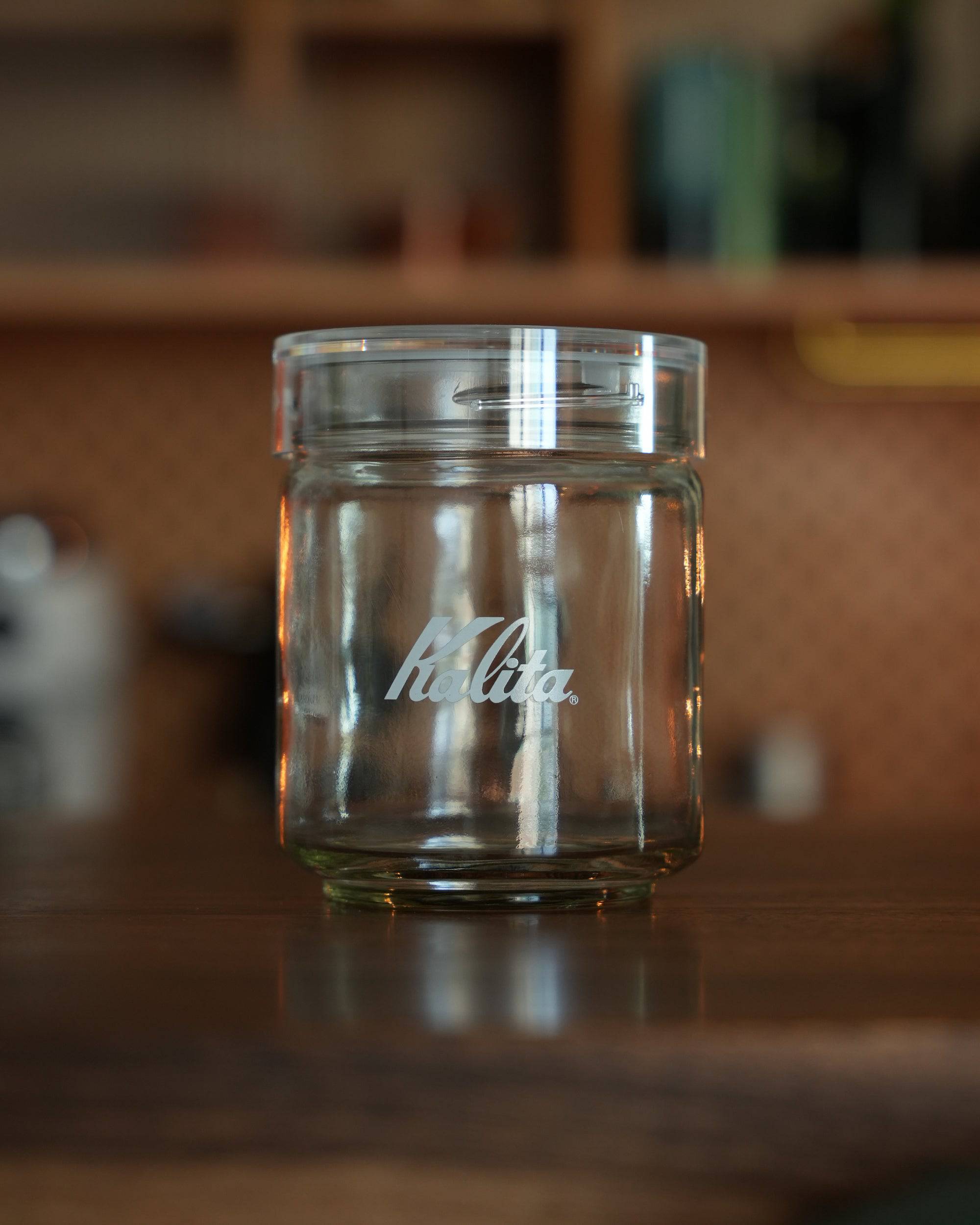 Transparent Kalita glass coffee storage jar with wide mouth and airtight lid, featuring brand logo, displayed on wooden surface in kitchen setting