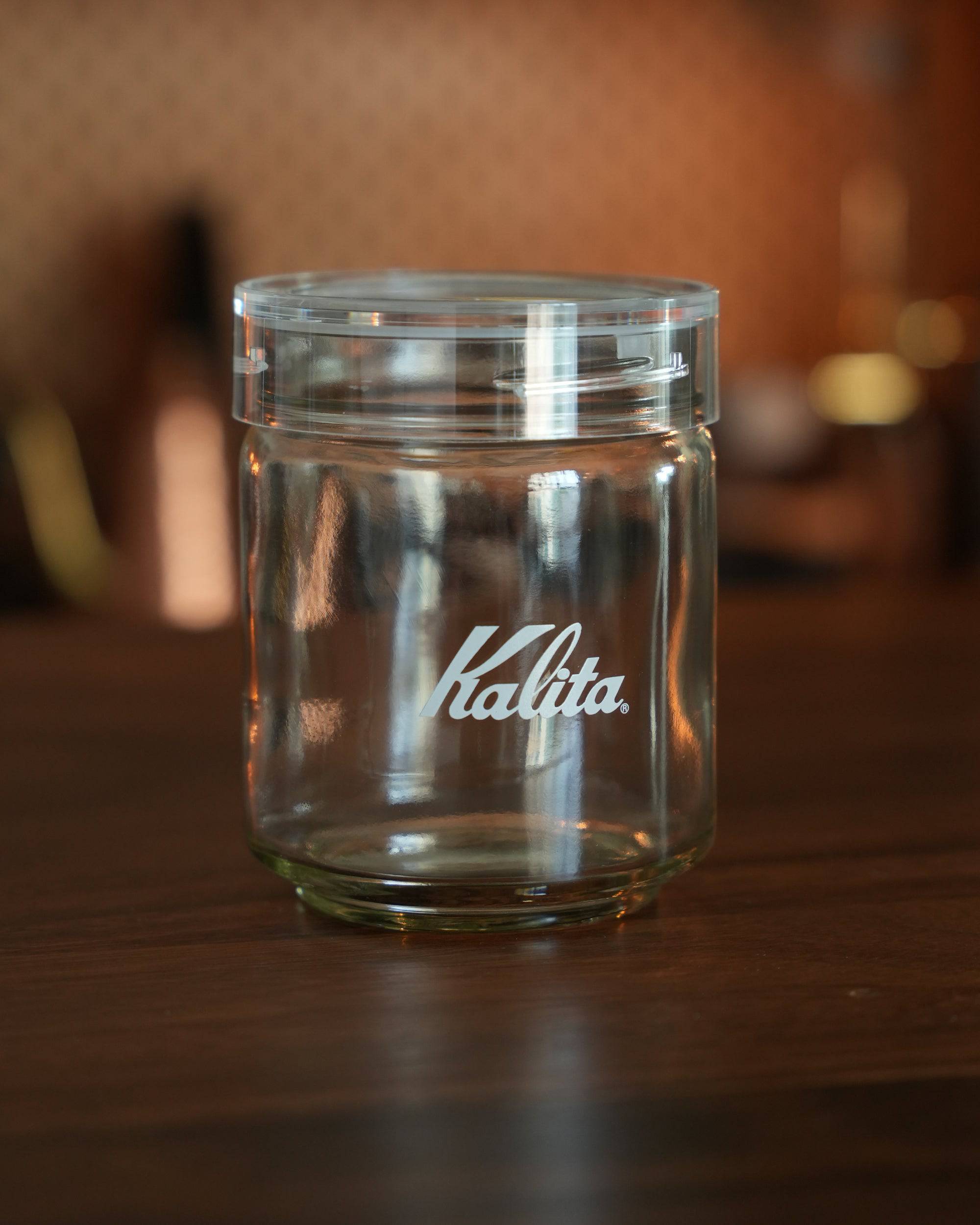 Kalita glass coffee storage jar with airtight lid, transparent design showcasing Kalita logo, placed on wooden surface with warm ambient lighting, ideal for preserving coffee beans freshness