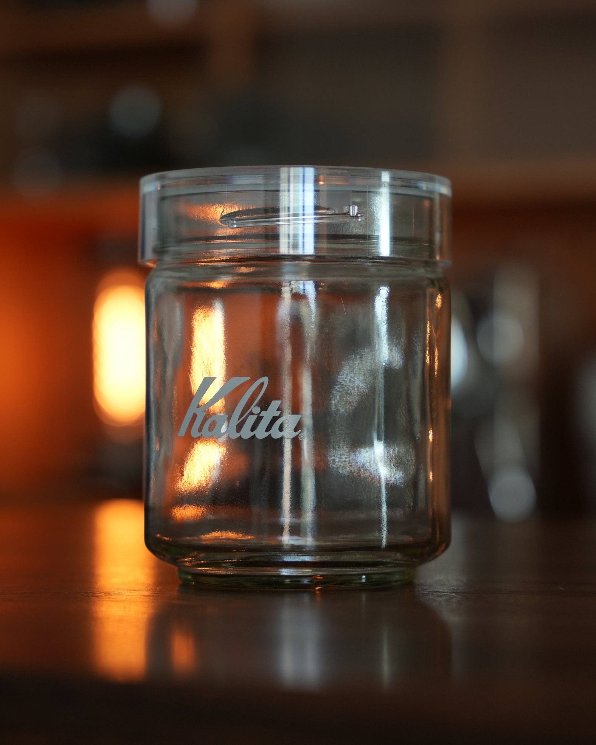 Kalita glass coffee storage jar with clear lid, displayed on wooden surface with warm ambient lighting, showcasing brand logo and airtight design for preserving coffee freshness