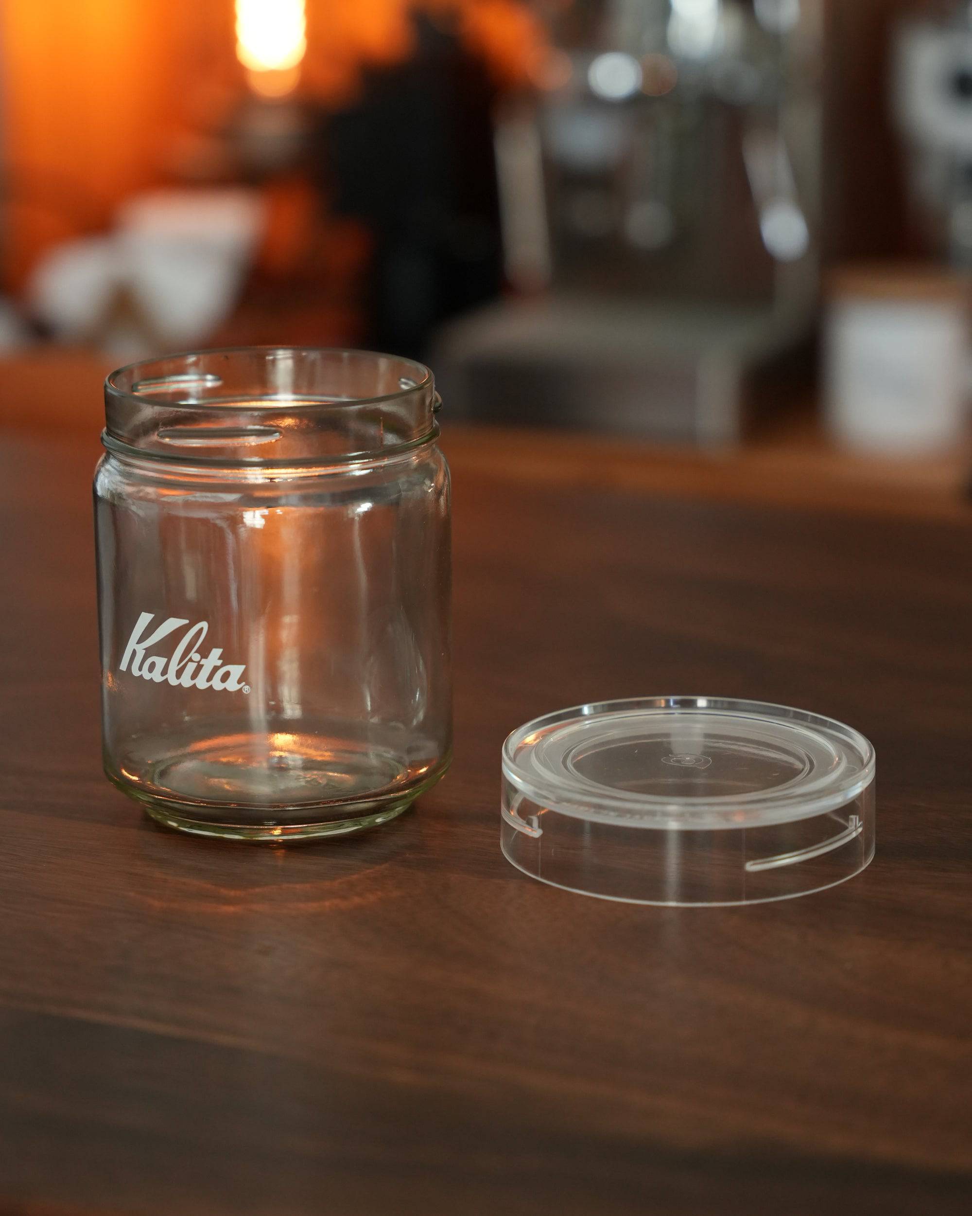 Kalita glass coffee storage jar with transparent lid on wooden table, showcasing branding and airtight seal design in warm cafe ambiance