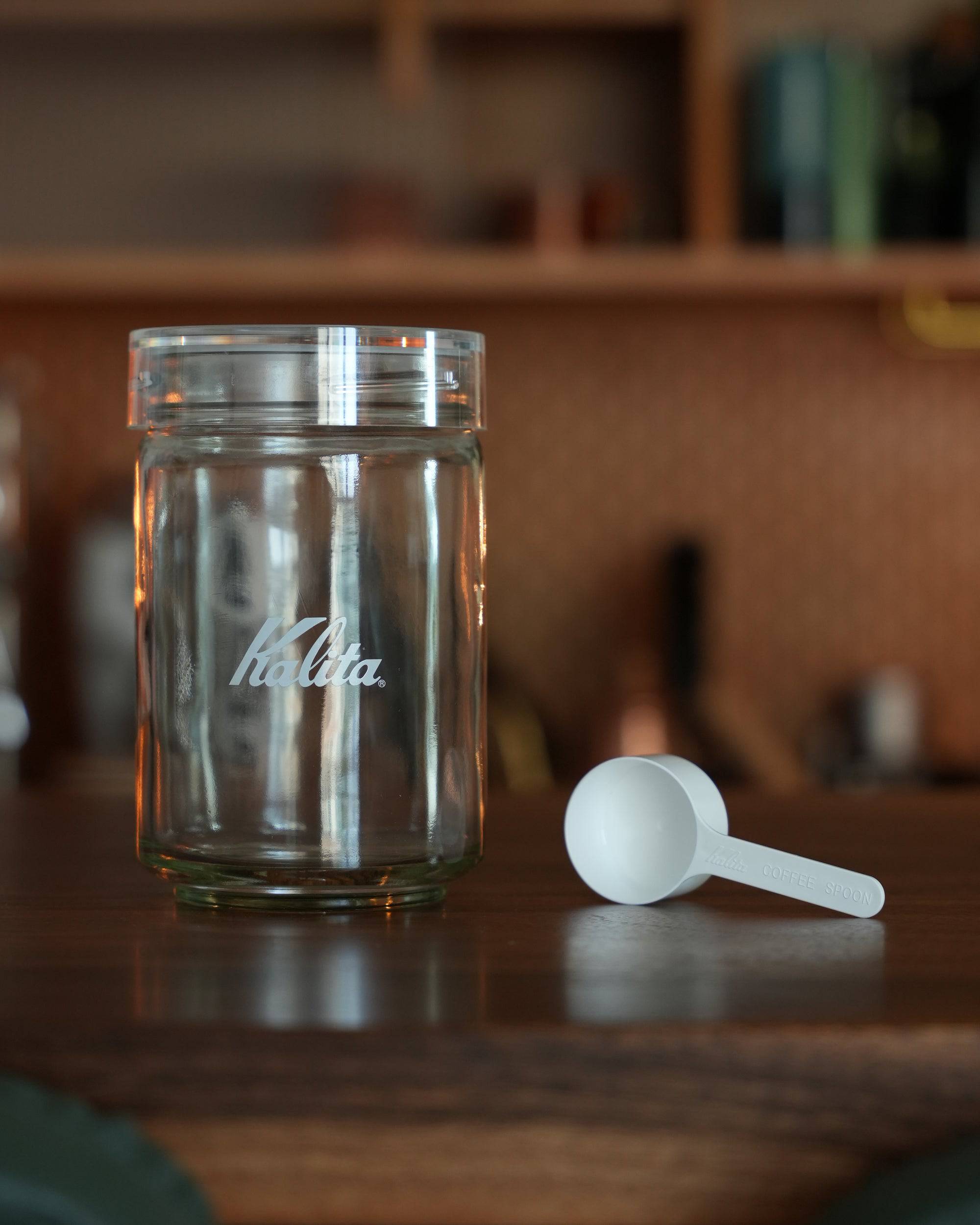 Kalita glass coffee canister with Kalita logo and white measuring scoop on wooden surface, kitchen background