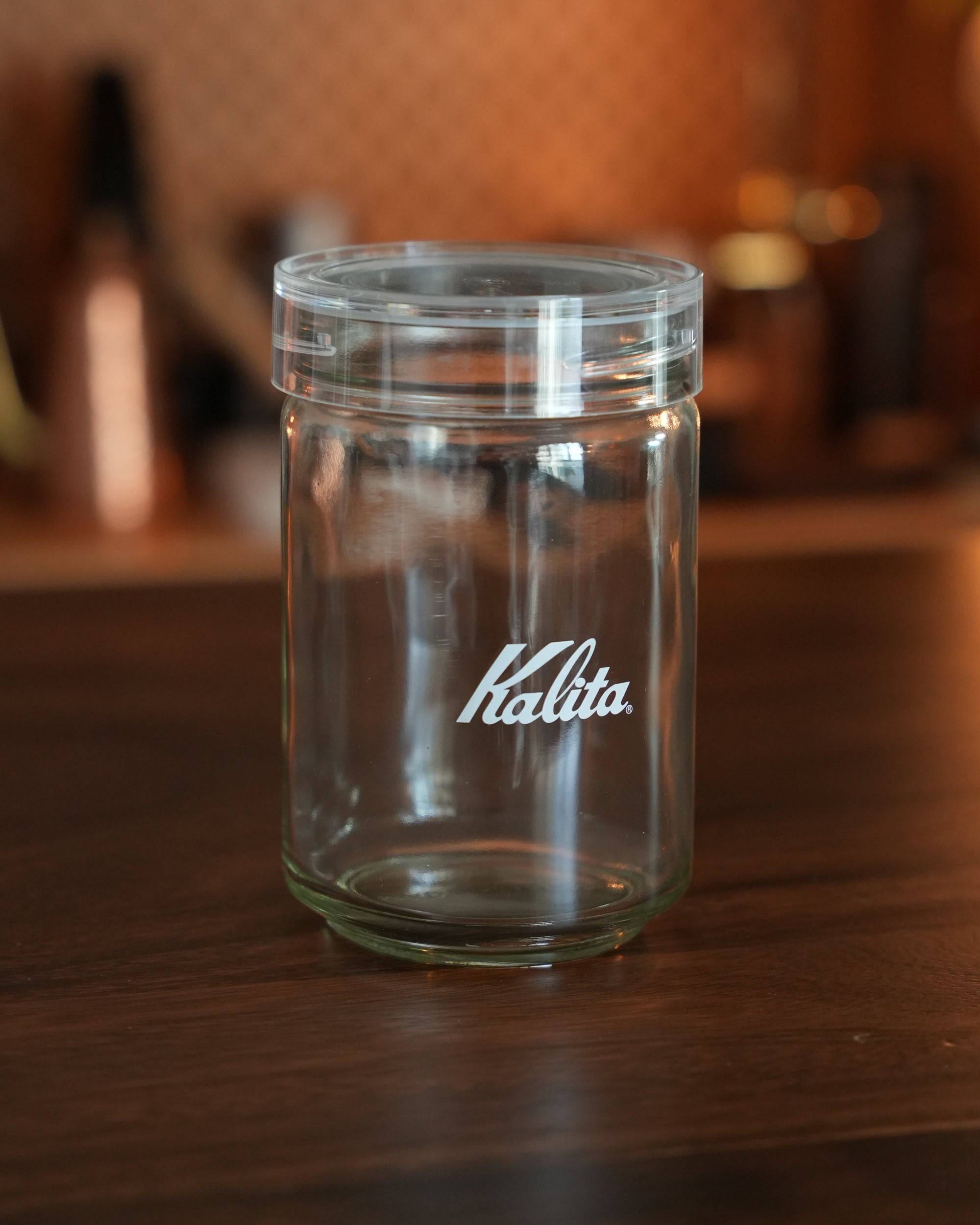 Kalita glass coffee storage jar with airtight lid, 300g capacity, clear container with brand logo, sitting on wooden surface with blurred background