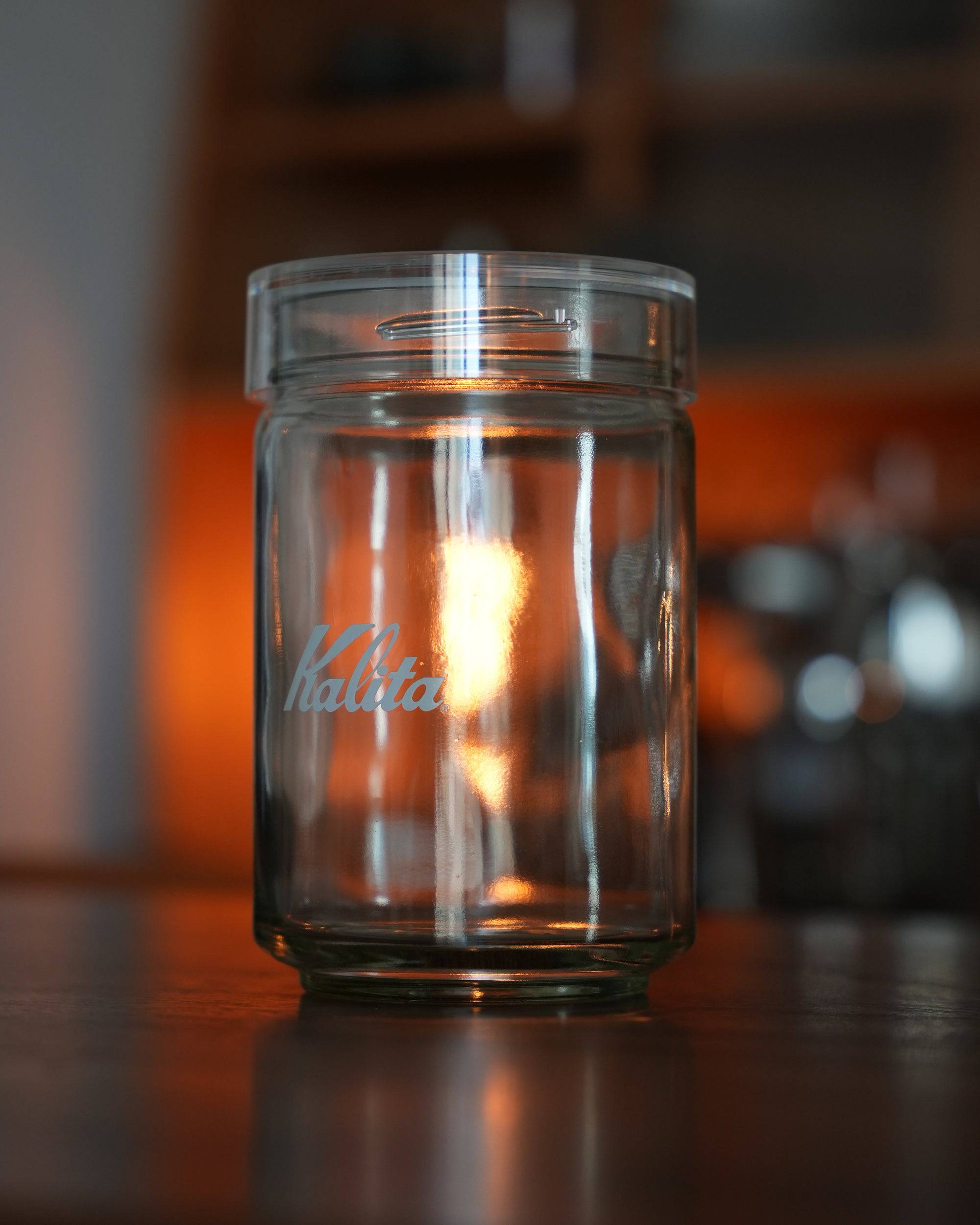 Kalita glass coffee canister with airtight lid, 300g capacity, showcasing brand logo, illuminated by warm ambient light, placed on wooden surface