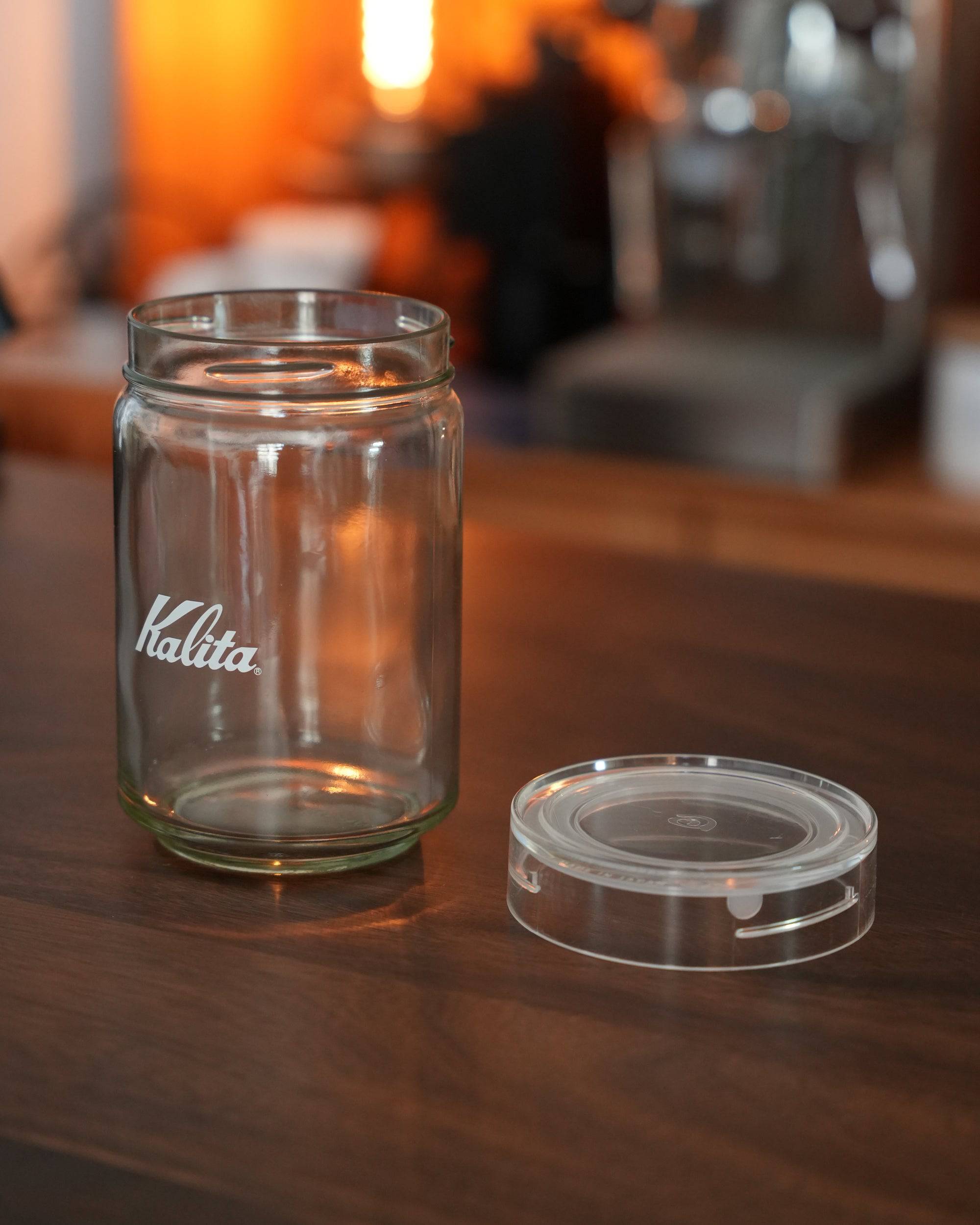 Kalita glass coffee storage jar with transparent lid on wooden surface, warm ambient lighting, blurred background with coffee equipment