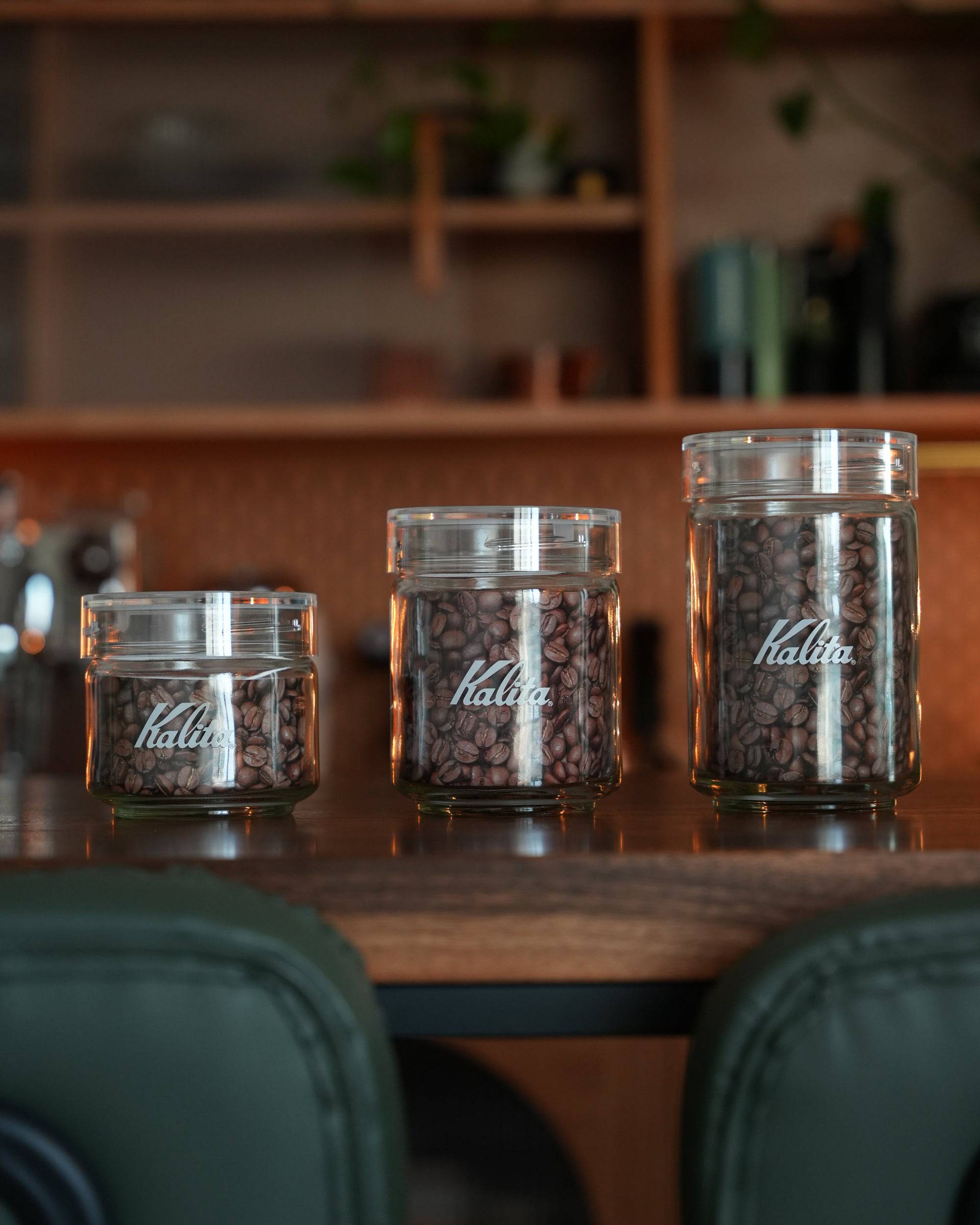 Three glass Kalita coffee storage jars of different sizes filled with coffee beans, displayed on a wooden counter with shelves in the background