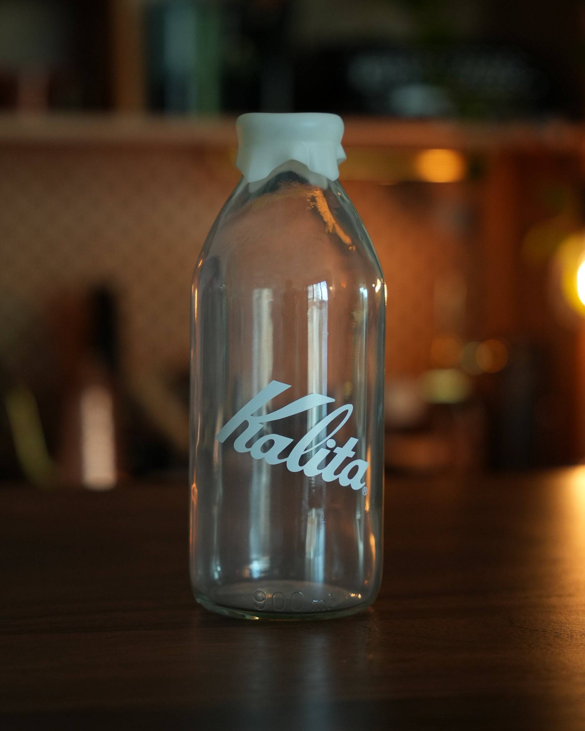 Kalita glass milk bottle shaped coffee bean storage jar, 320g capacity, with white lid and blue logo, on wooden surface in warm-lit kitchen setting