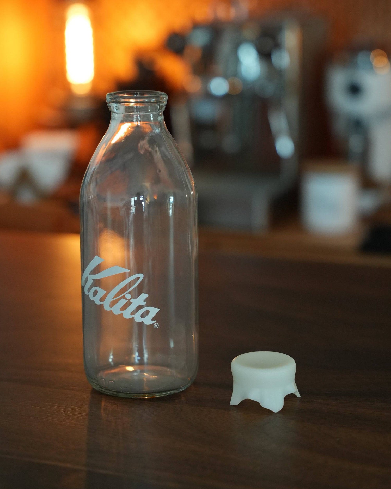 Kalita glass milk bottle coffee bean storage jar on wooden counter with espresso machine in background, warm lighting ambiance