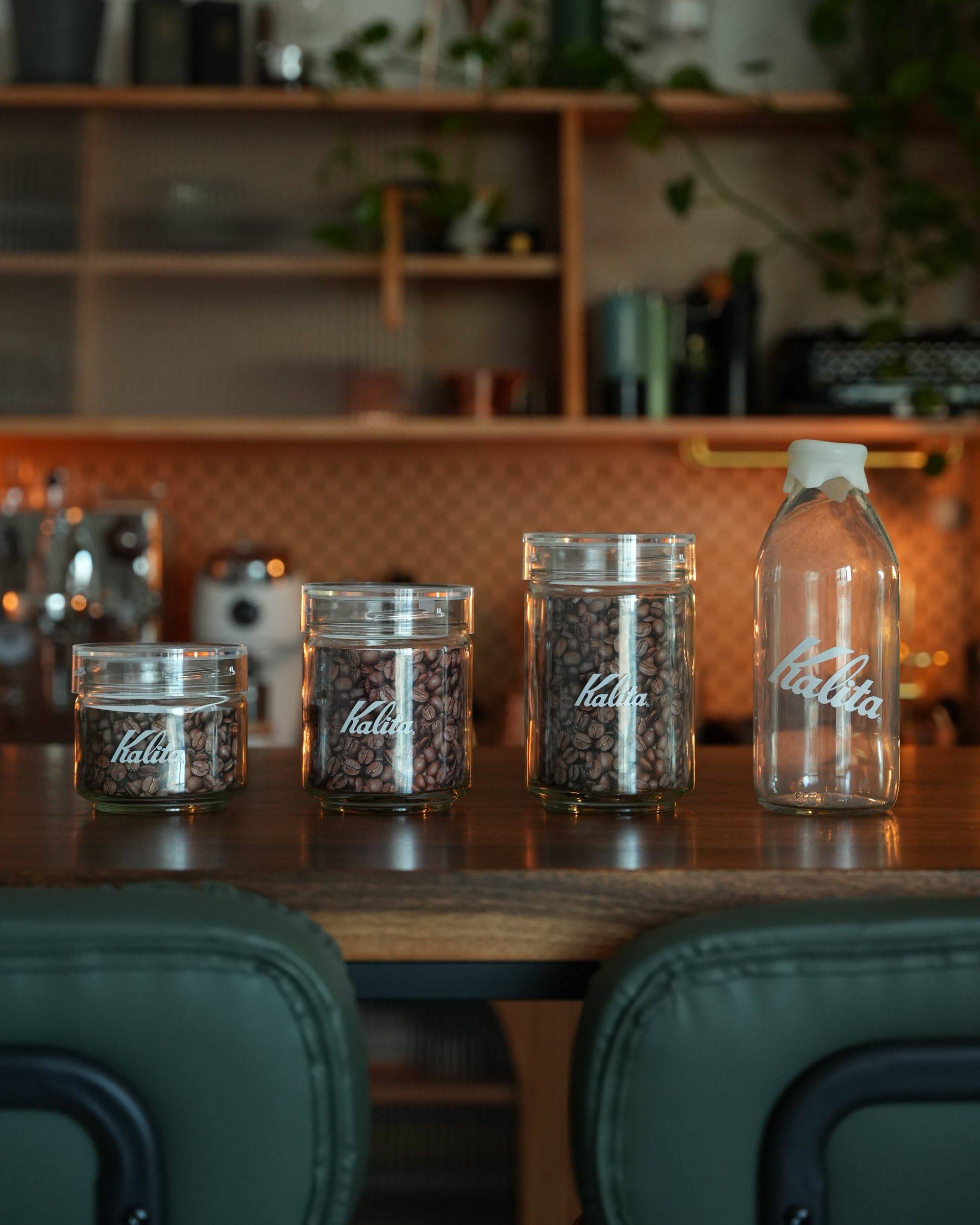 Kalita glass coffee bean storage jars in various sizes displayed on kitchen counter, ranging from small to large milk bottle shape, filled with coffee beans, showcasing stylish design and functionality