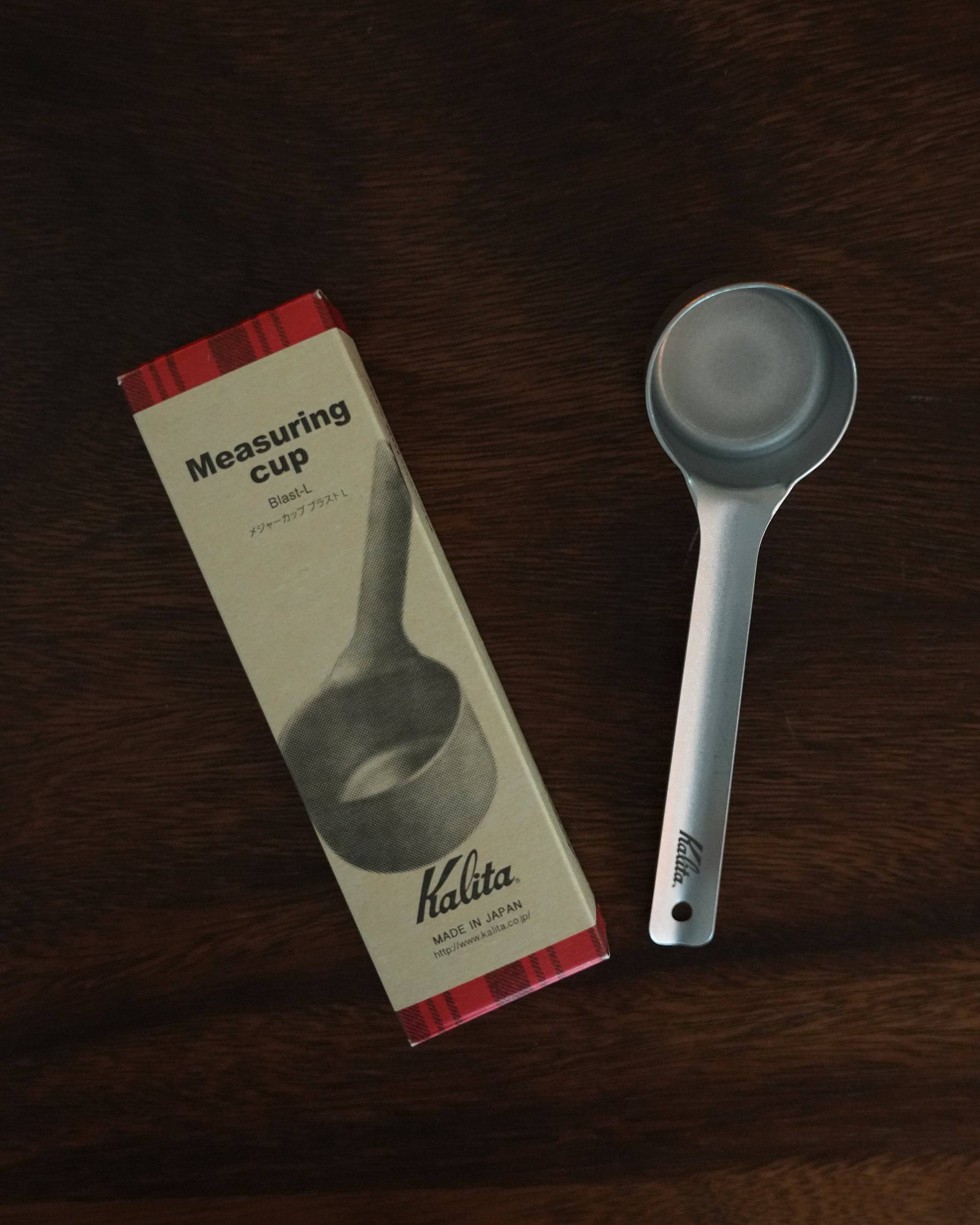 Kalita stainless steel coffee measuring spoon 10g (L Size) displayed next to its packaging on a dark wooden surface, showcasing the product&