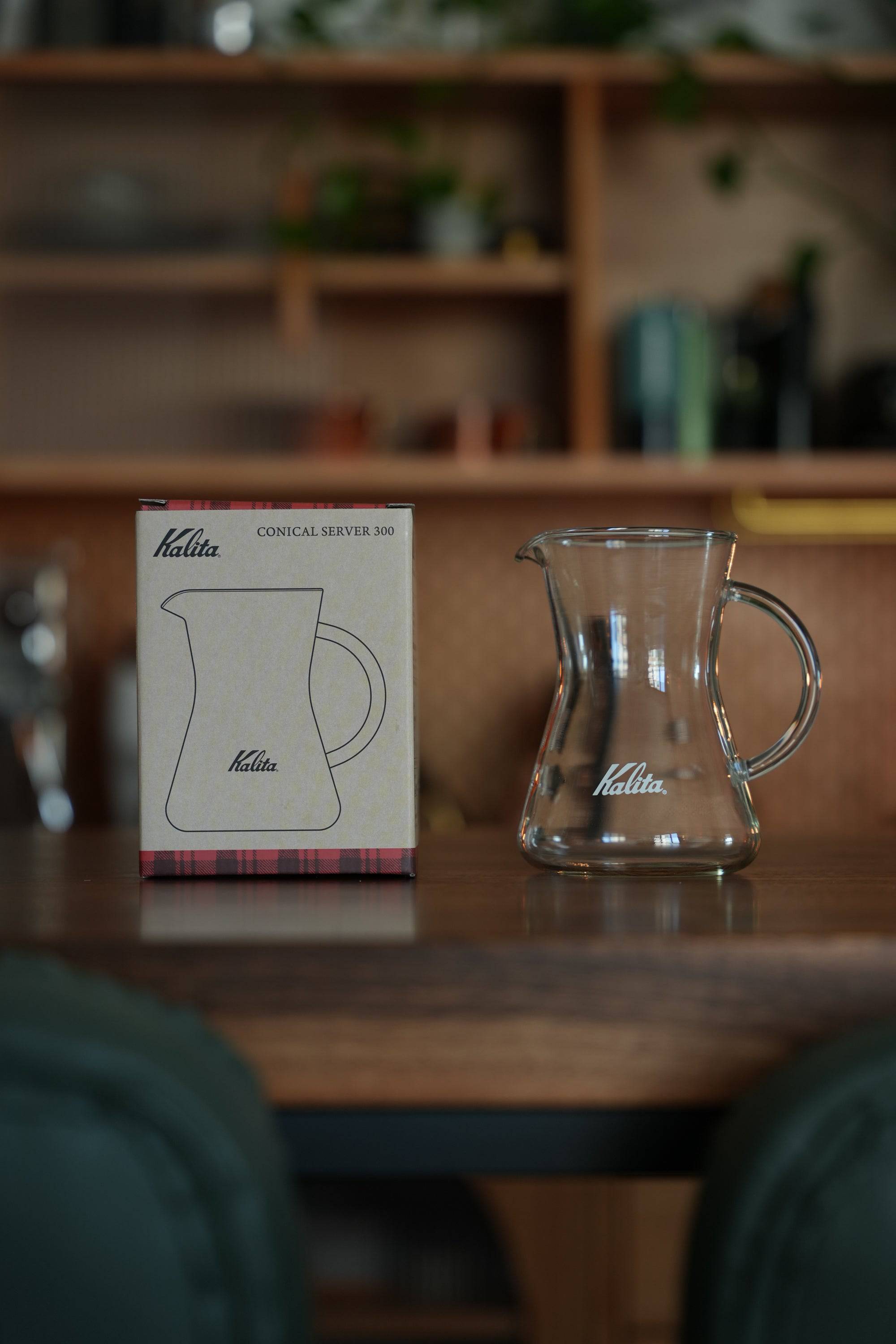 Kalita conical server 300 glass coffee carafe displayed with its packaging box on wooden counter, showcasing elegant design and heat-resistant glass construction for stylish coffee brewing and serving