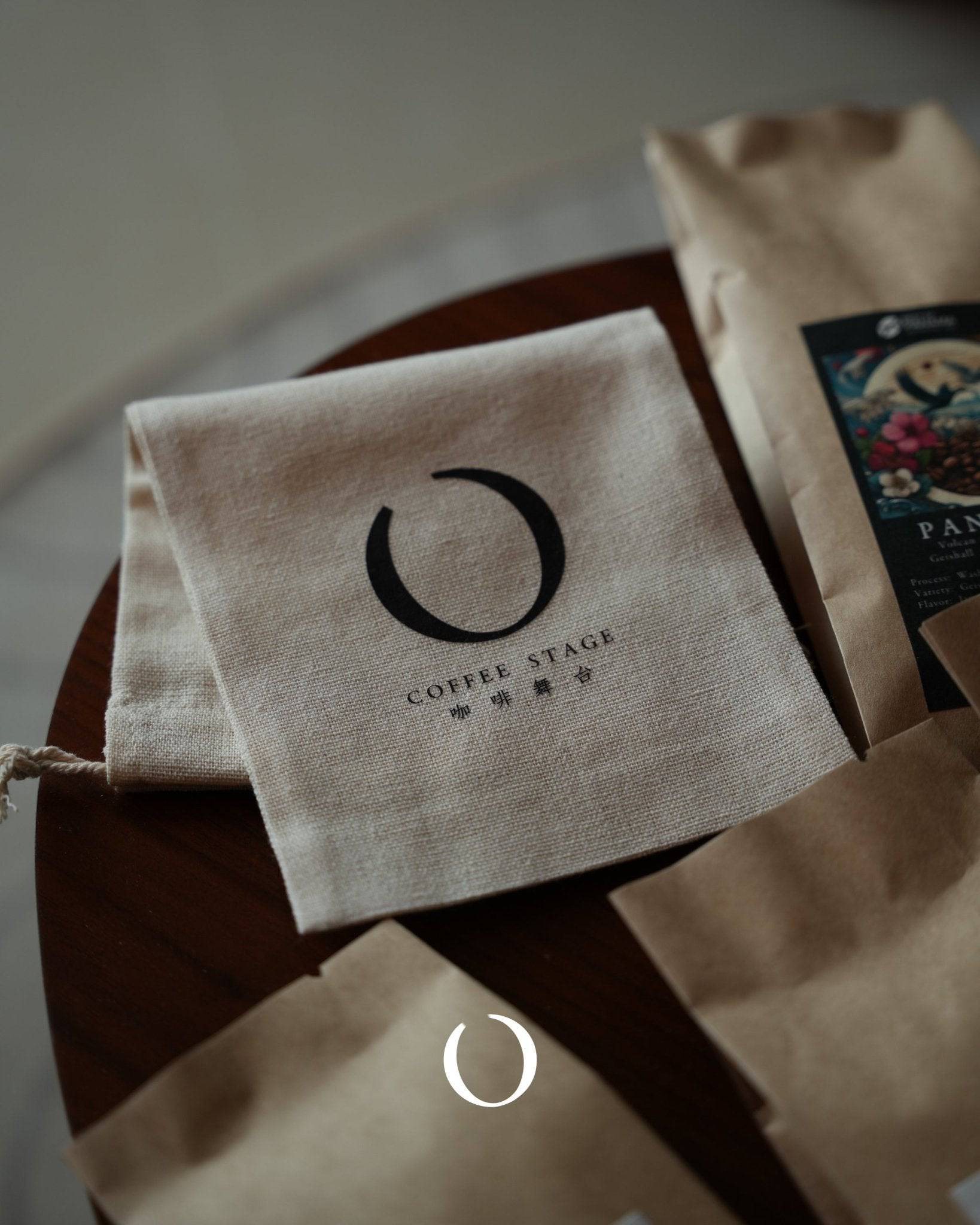 Coffee packaging from Coffee Stage, featuring a cream-colored cloth napkin with logo and brown paper bags on a wooden surface, showcasing premium Ethiopian Ana Sora coffee products