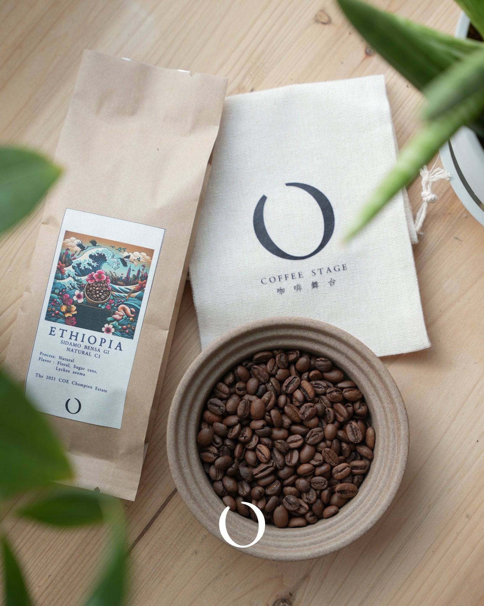 Ethiopian coffee beans from Bensa Coffee estate displayed with packaging, a ceramic bowl of roasted beans, and a cloth bag featuring the Coffee Stage logo, set on a wooden surface with plant leaves in frame
