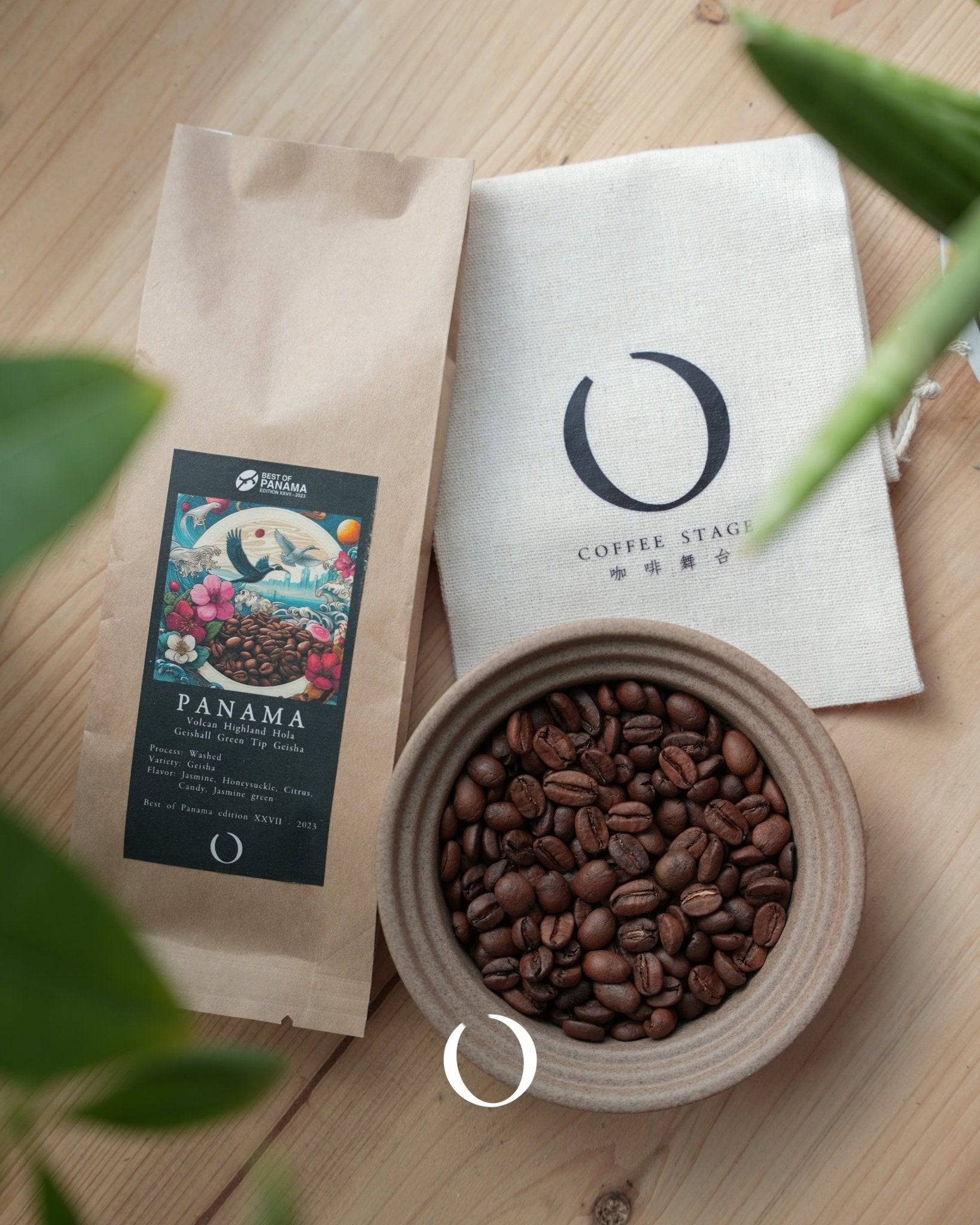 Premium Panama Geisha coffee beans in a ceramic bowl, alongside branded packaging and a Coffee Stage cloth, on a wooden surface with green leaves in view
