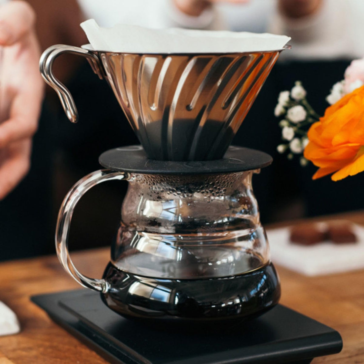 HARIO V60 02 silver stainless steel dripper, 1–4 cups: The V60 02 dripper is brewing over a glass server; the gooseneck kettle, glass server and scale are scene props.