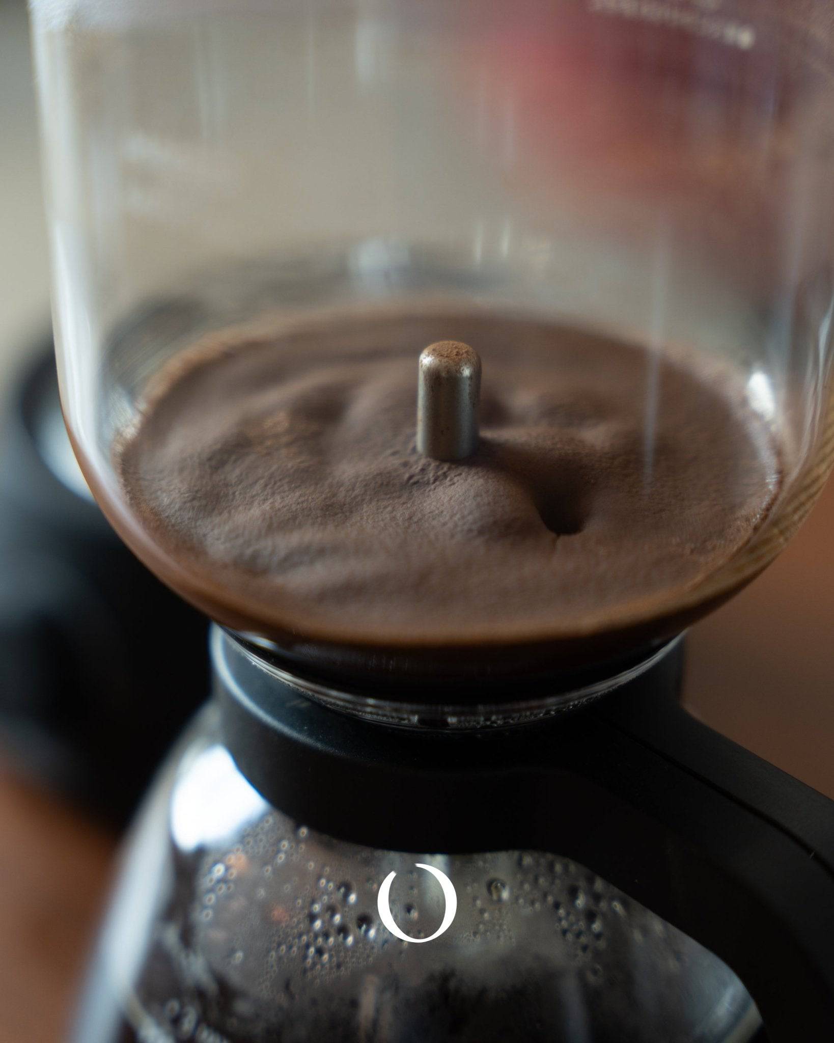 Closeup of HARIO electric siphon coffee maker brewing process, showing coffee grounds in upper chamber and bubbling water in lower chamber, demonstrating the unique vacuum brewing method for rich, flavorful coffee extraction.