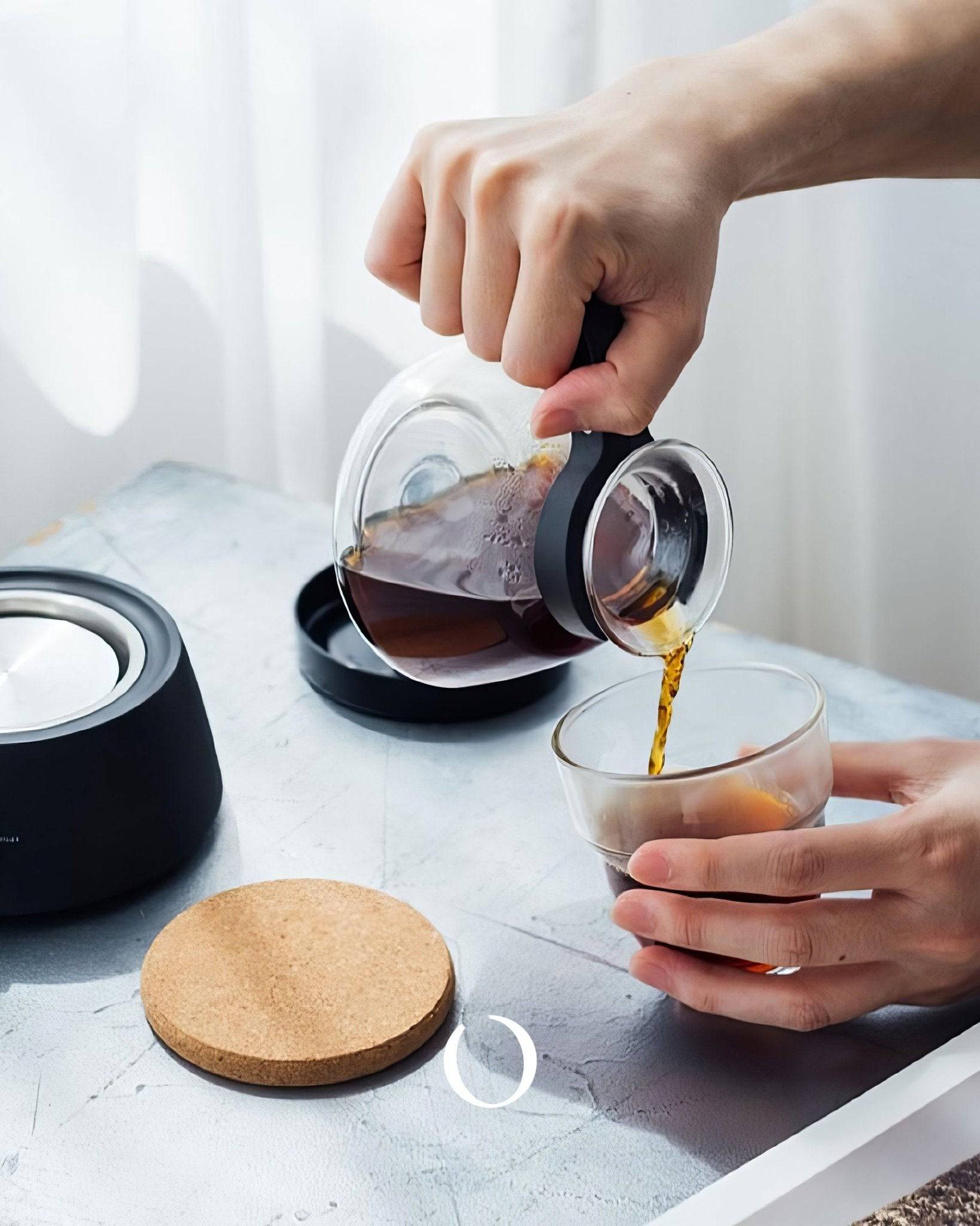 Hand pouring fresh brewed coffee from HARIO electric siphon coffee maker into glass cup, with black base unit and cork coaster visible on marble countertop