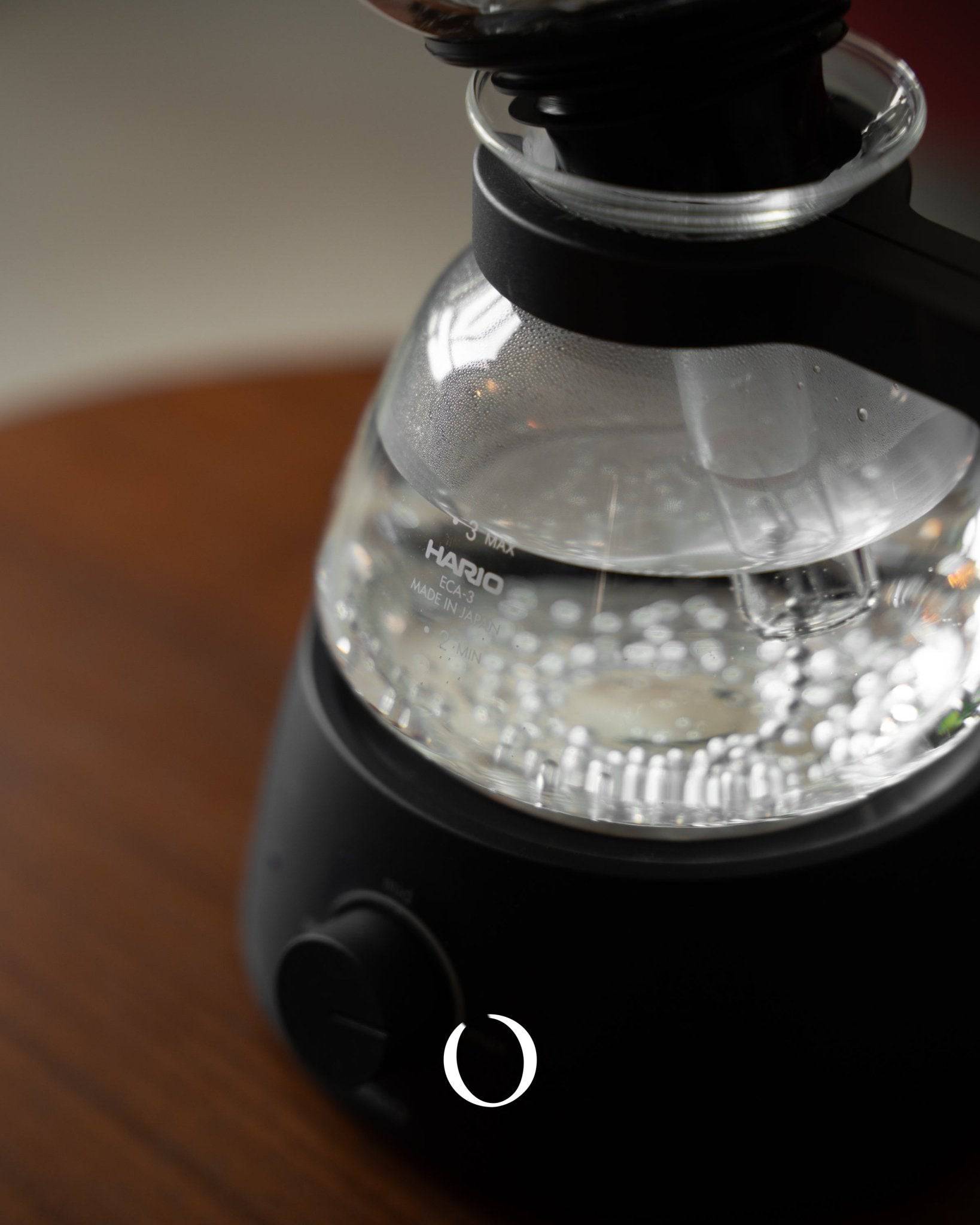 HARIO electric siphon coffee maker with glass upper chamber and black base, boiling water visible inside, showcasing unique brewing process and elegant design for coffee enthusiasts