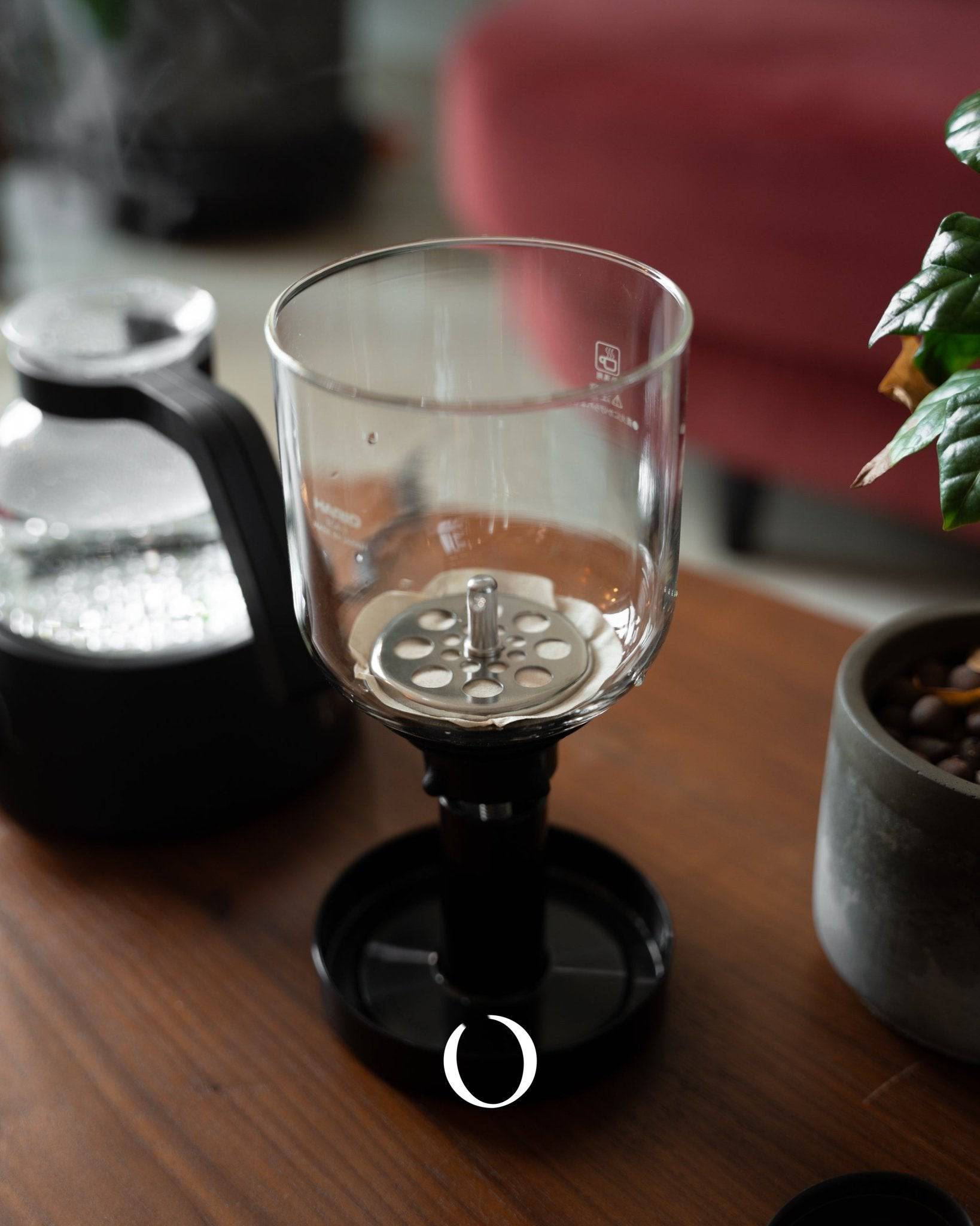 HARIO electric siphon coffee maker ECA-3-B with glass upper chamber and metal filter on wooden table, next to black base unit and potted plant, showcasing elegant design and brewing process