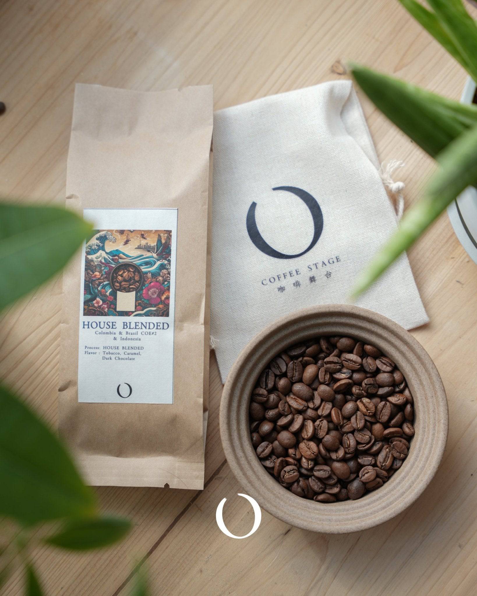House Blended coffee packaging with colorful label, ceramic bowl of roasted coffee beans, and branded cloth bag on wooden surface, surrounded by green plant leaves