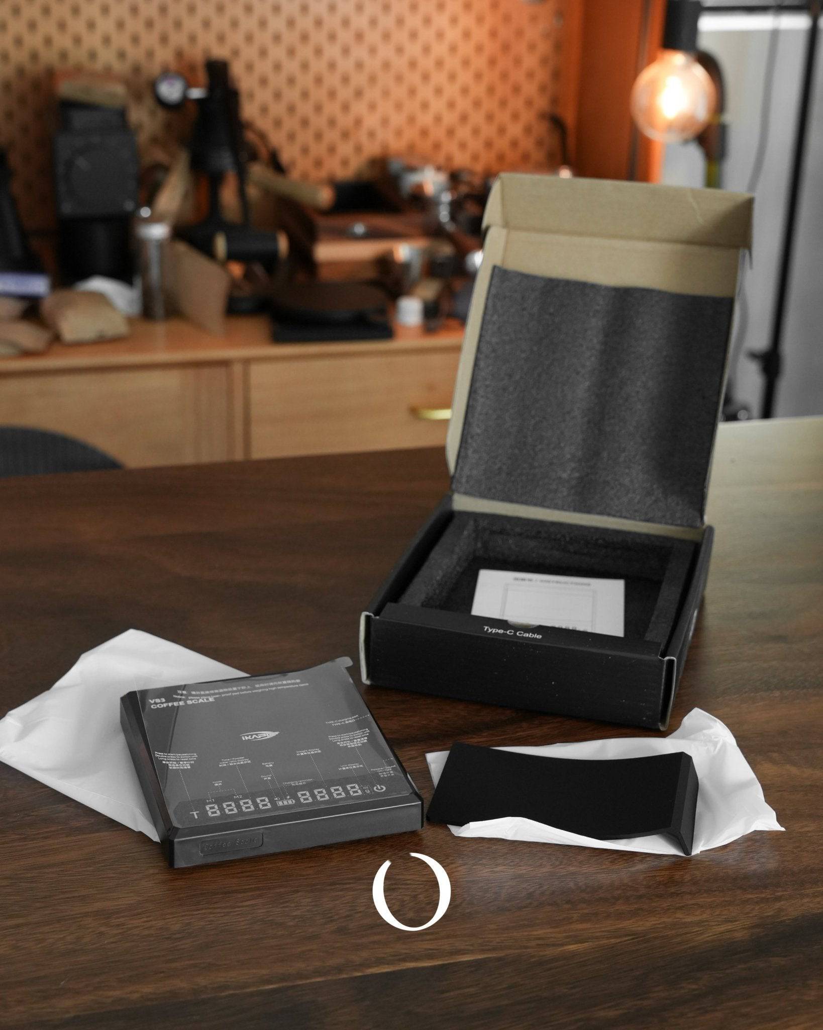 Ikape Espresso Black Warrior coffee digital scale with packaging, display screen, and accessories on wooden table in office setting. High-precision scale for brewing perfect espresso and pour-over coffee.