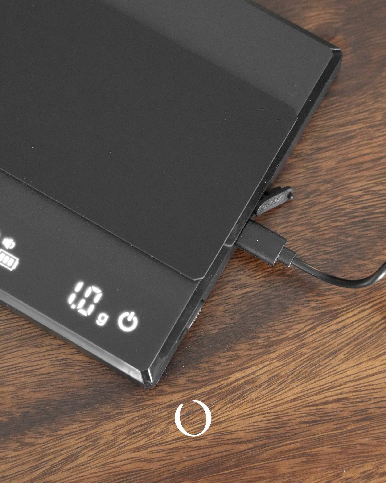 Sleek black Ikape Espresso digital coffee scale with LED display showing 1.0g, charging cable connected, on wooden surface. Precision weighing tool for coffee enthusiasts.