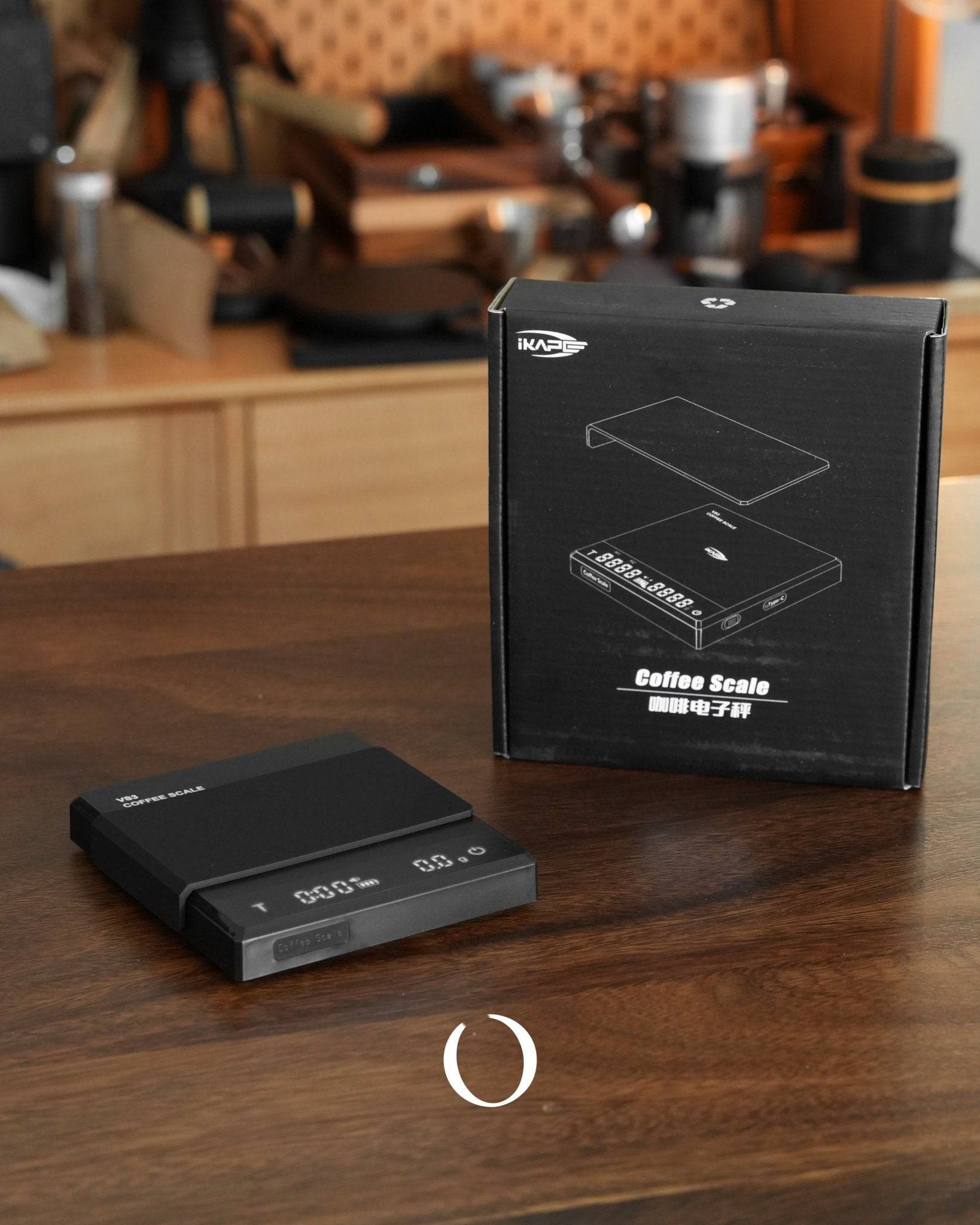 Ikape Espresso Black Knight coffee scale with product box on wooden table, surrounded by coffee equipment in background. Sleek black design with digital display, showcasing precision and functionality for coffee enthusiasts.