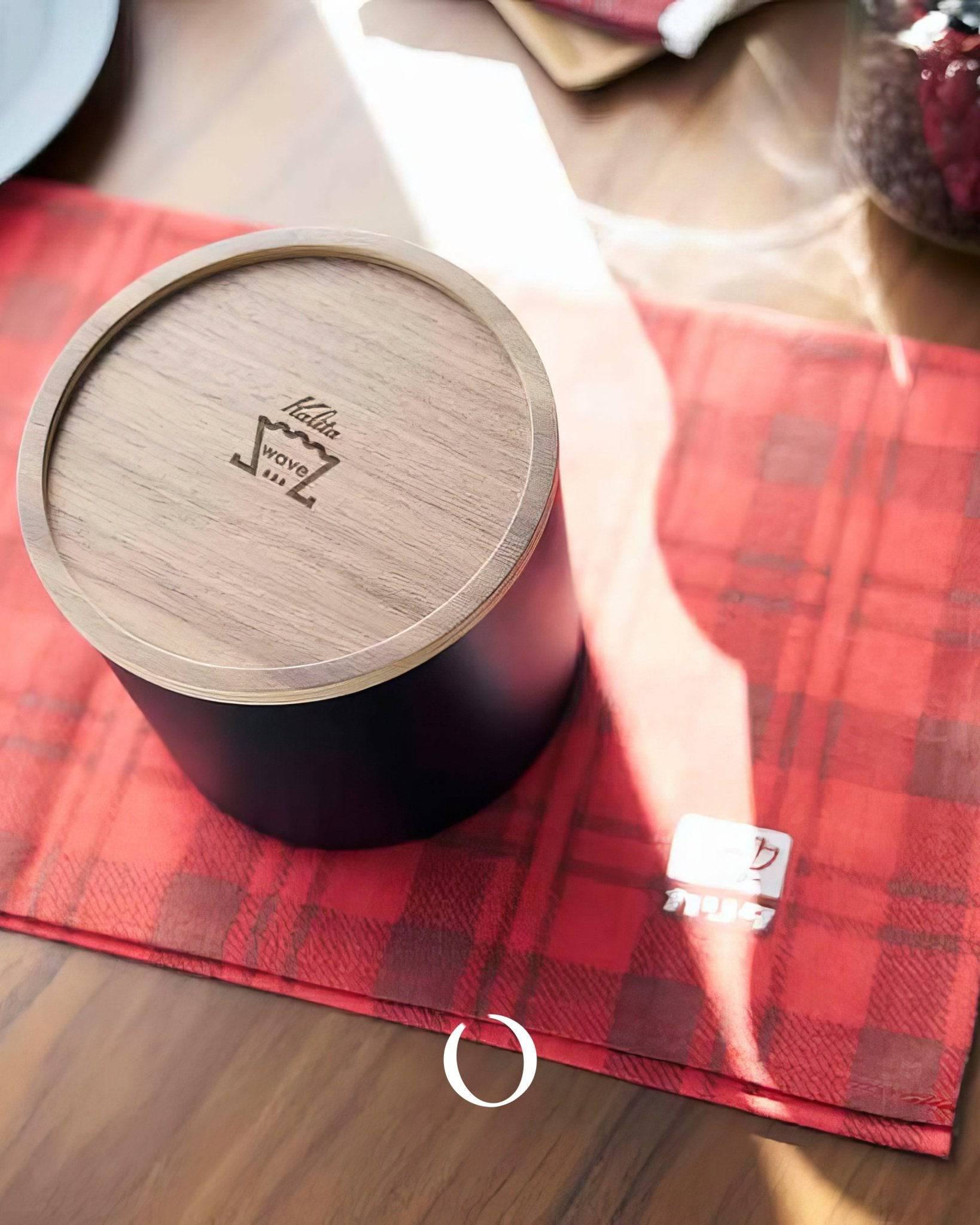 Kalita 155/185 Roshi filter paper container on red plaid napkin, wooden lid with logo visible, sunlight casting shadows, coffee accessories on table