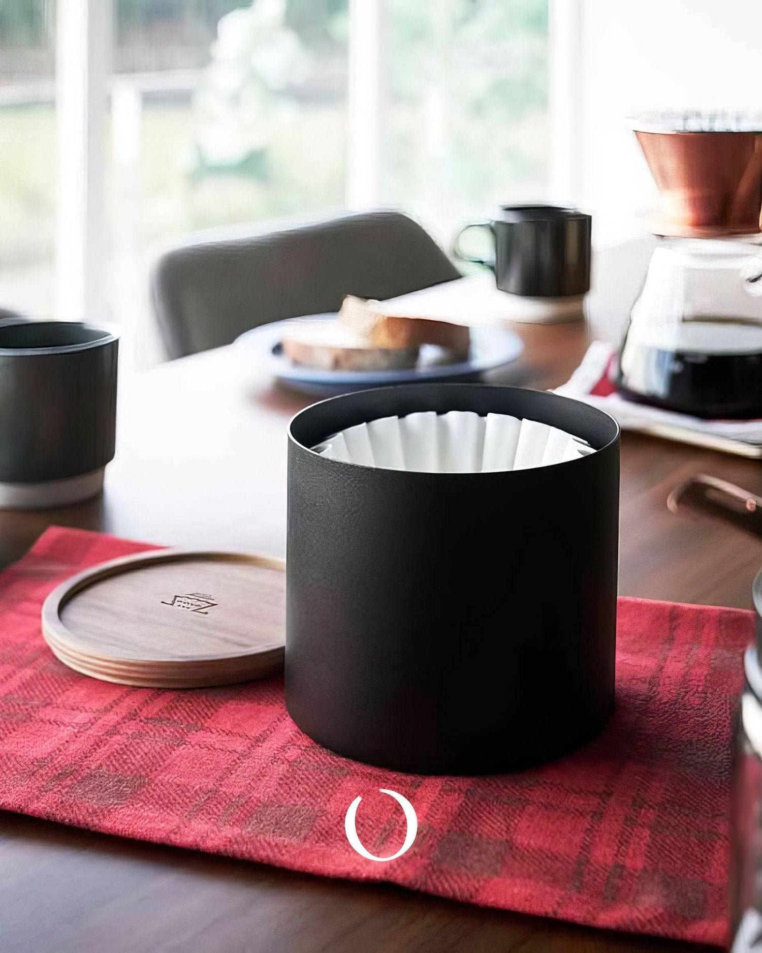 Kalita 155/185 Roshi filter paper canister on red plaid cloth, wooden table with coffee setup, mugs, and breakfast plate in background, showcasing stylish coffee accessories for home brewing