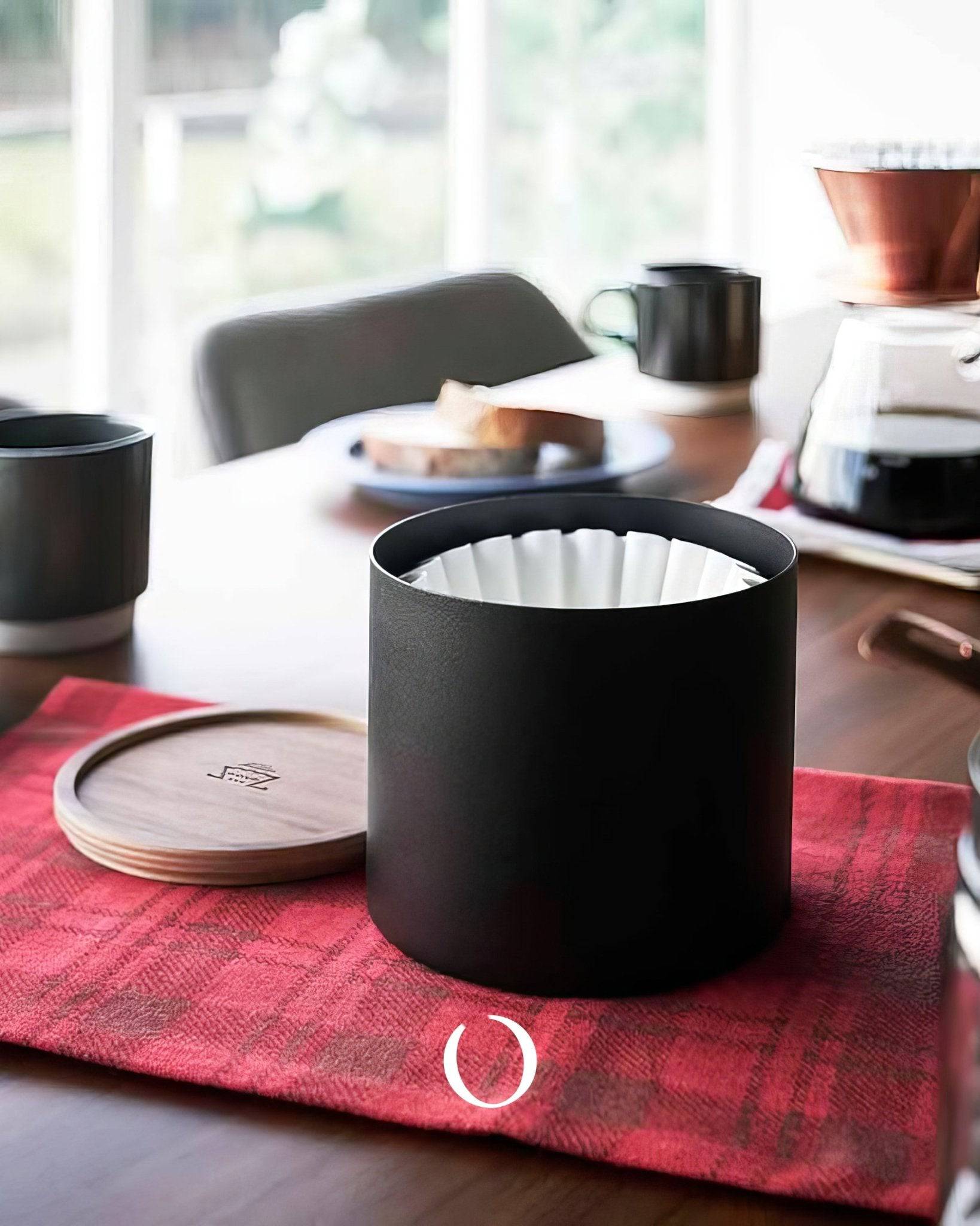 Kalita 155/185 Roshi filter paper canister on red plaid cloth, wooden table with coffee setup, mugs, and breakfast plate in background, showcasing stylish coffee accessories for home brewing