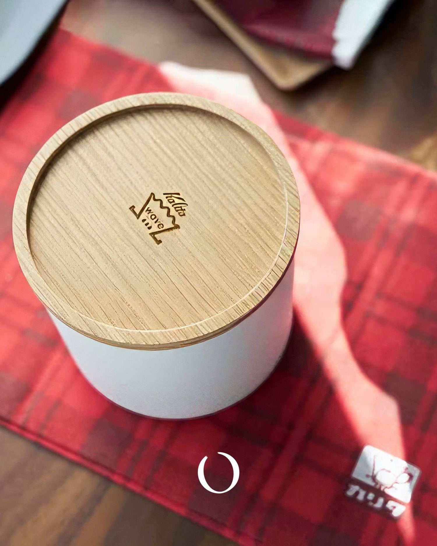 Kalita 155/185 Roshi filter paper container with wooden lid on red plaid cloth, showcasing elegant design for coffee enthusiasts. White cylindrical body with Kalita logo engraved on top, perfect for storing and protecting coffee filters.