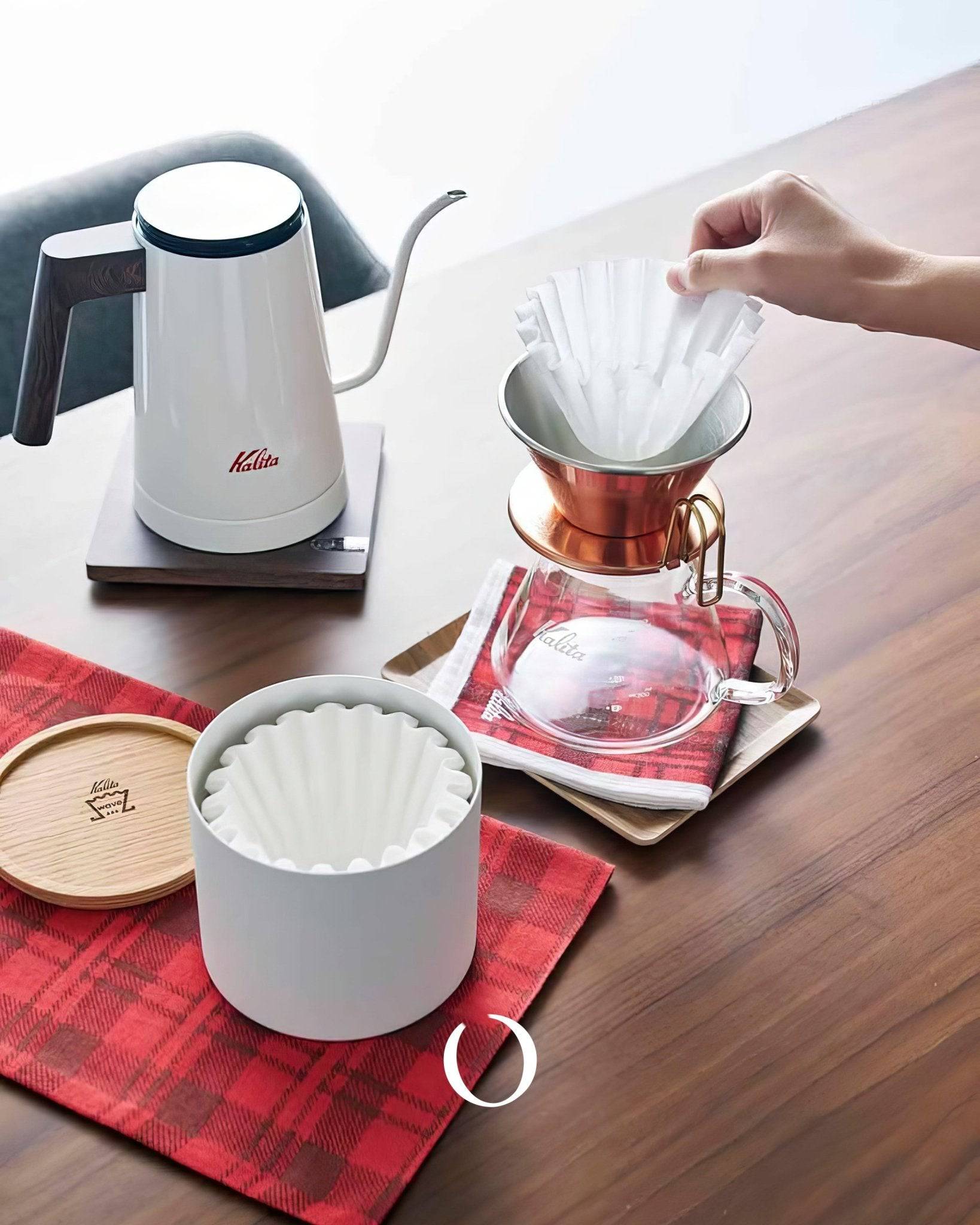 Kalita coffee brewing set with electric kettle, pour-over dripper, white filter canister, and red plaid cloth on wooden table. Hand placing filter in copper dripper.