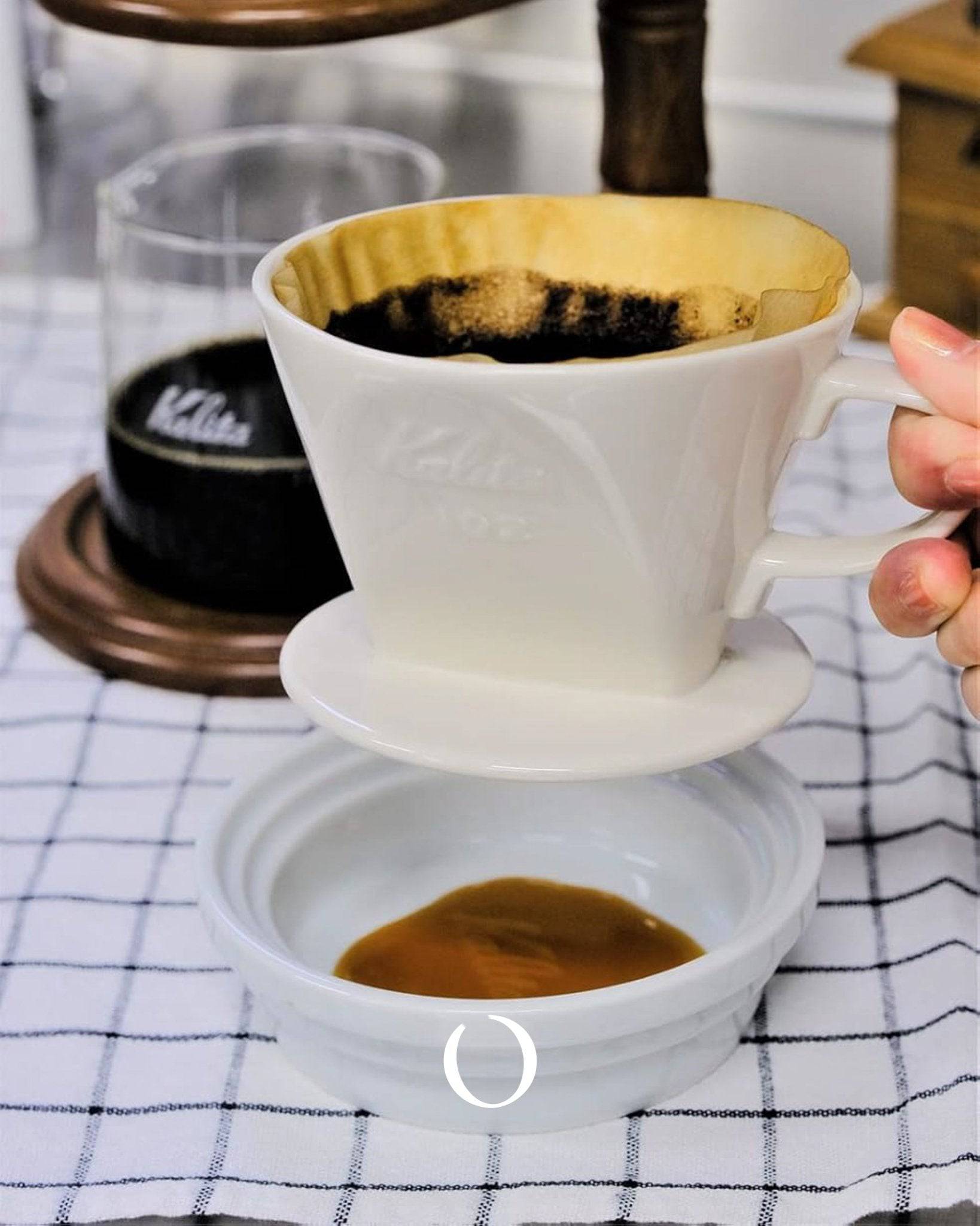 Kalita & Hasami ceramic coffee dripper and saucer set in white, showcasing Japanese craftsmanship for pour-over brewing, placed on a checkered cloth with coffee being prepared