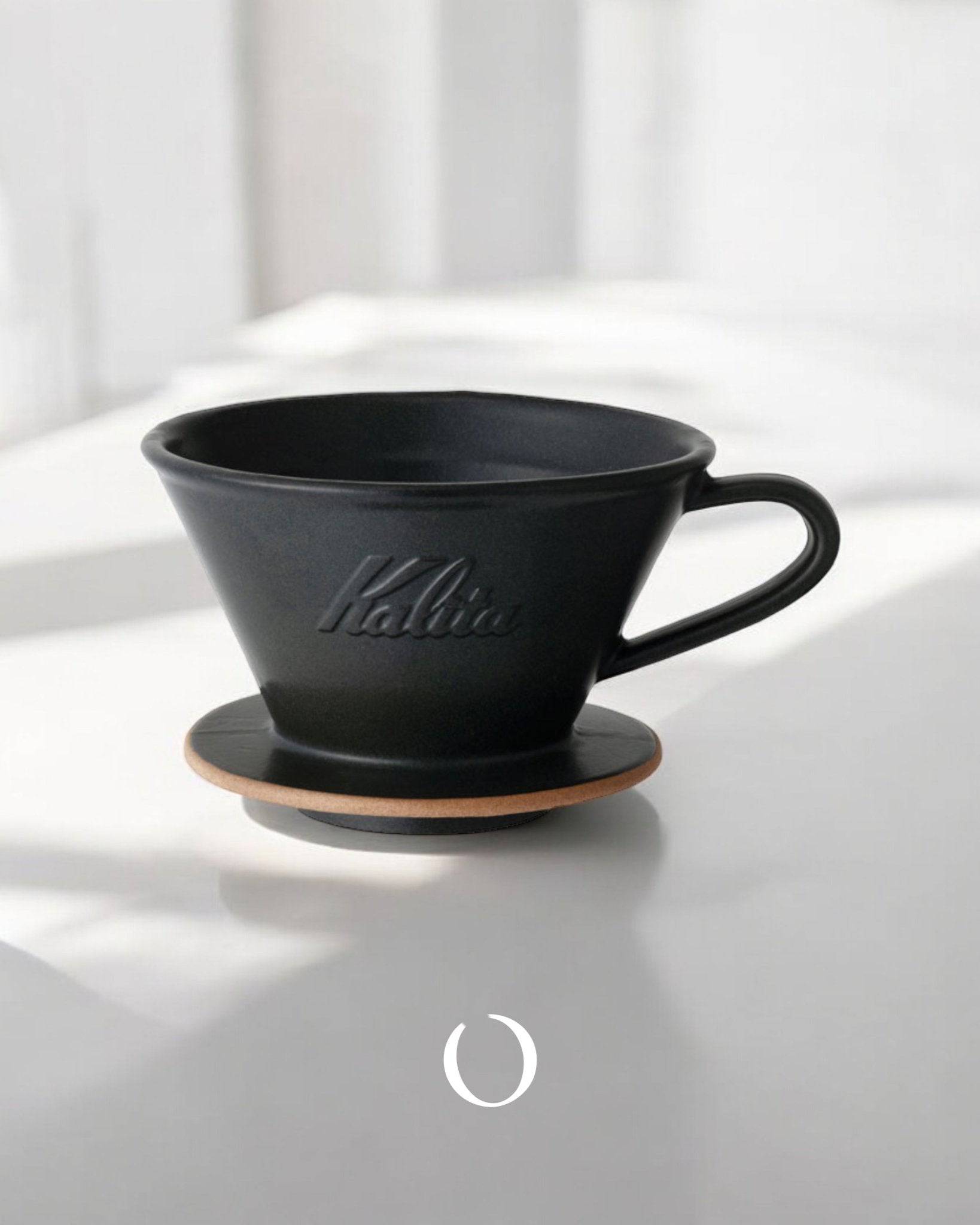 Kalita MI185 ceramic pour-over coffee dripper in matte black, featuring a wide base and Kalita branding, placed on a wooden coaster against a white background with soft shadows