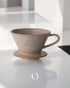 Kalita MI185 ceramic pour-over coffee dripper in beige color, featuring a conical shape with wide rim, handle, and branded logo, positioned on a white surface in a minimalist setting